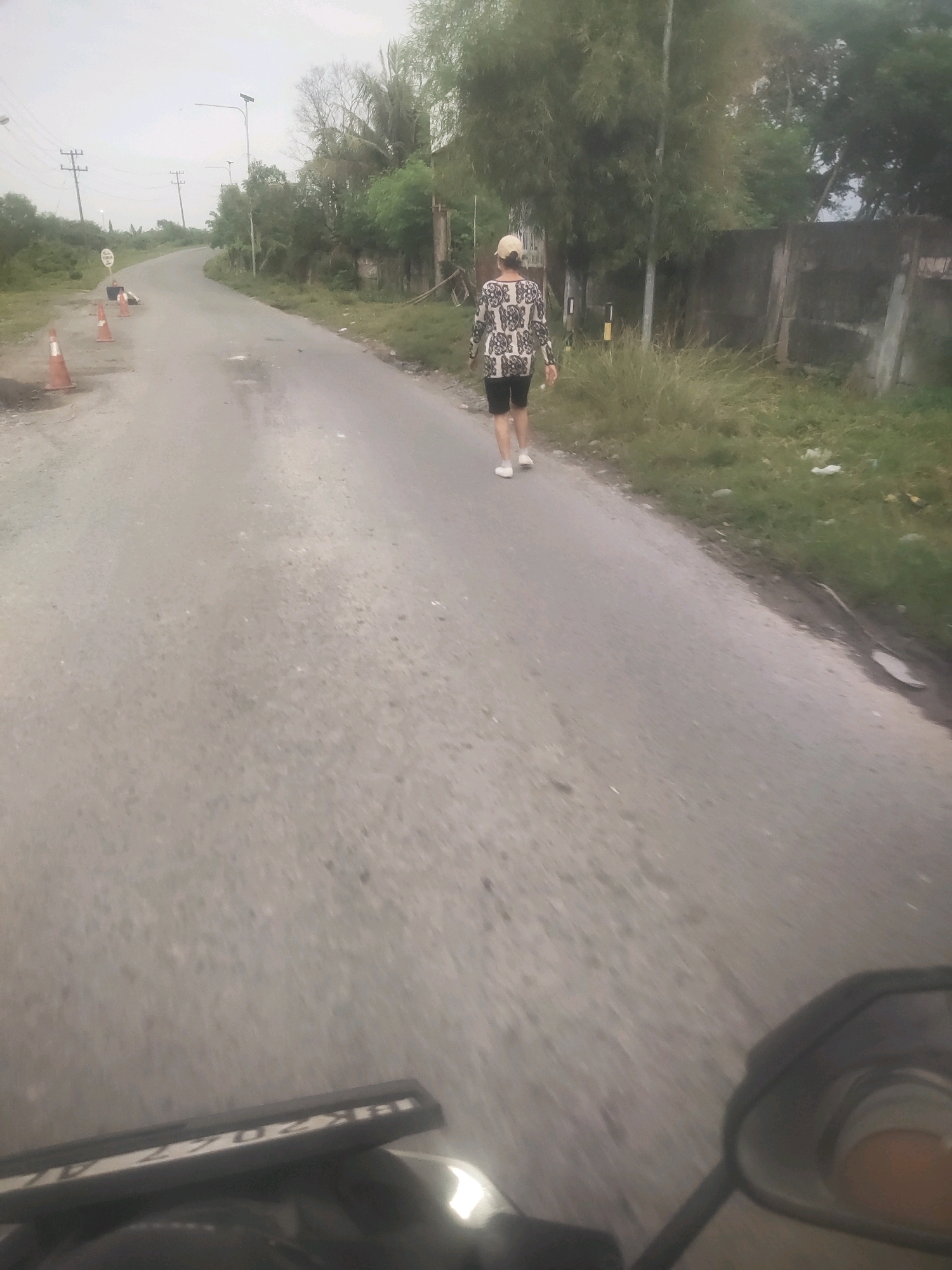 A person wearing a patterned long-sleeved shirt, black shorts, and a light-colored baseball cap walks away from the camera on a paved road. The road curves to the left in the background. To the right of the road is a low concrete wall covered in moss and greenery, with trees and bamboo behind it. To the left of the road, there are traffic cones, power lines with utility poles, and some vegetation. The sky is overcast. The bottom of the image shows a portion of a motorcycle, including its license plate and a mirror. The license plate appears to read "BK 2967 AL". The scene is likely in Medan, Indonesia, given the license plate format and tropical vegetation.