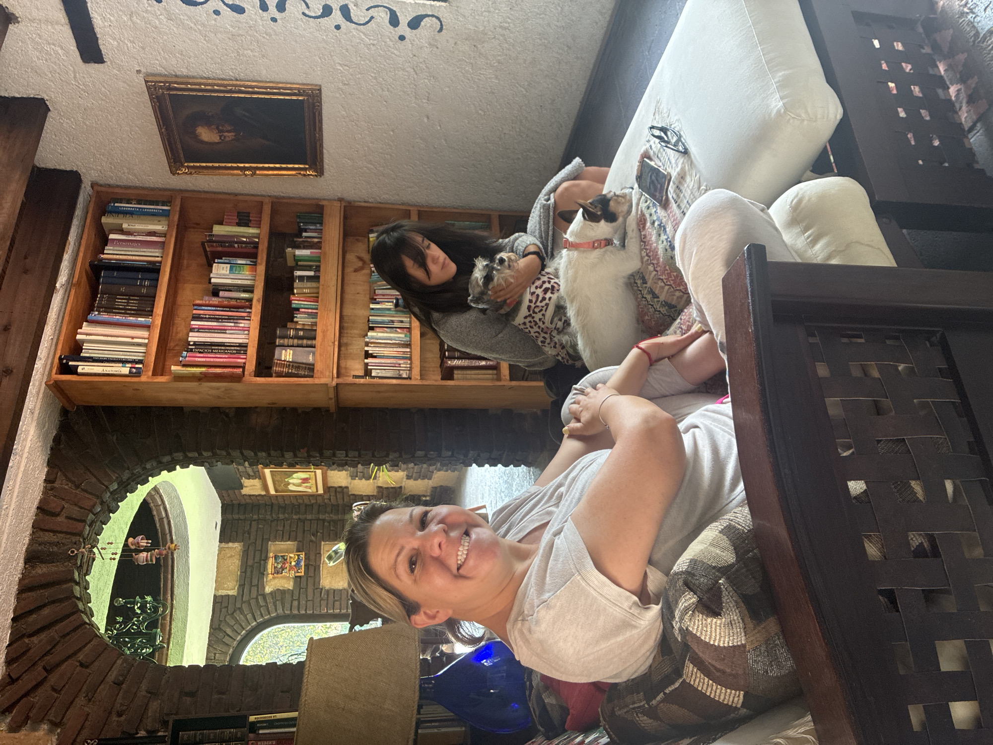 A bright, naturally-lit interior shows two women on a light-colored couch. One woman is facing the camera and smiling broadly, she's reclined, wearing a light-colored t-shirt and patterned shorts. The second woman, facing away from the camera, is seated and holds a small dog wearing a patterned shirt. A larger white dog with a pink collar stands next to her. Behind the women, a bookshelf filled with books and a framed picture are visible. A stone archway is seen behind the woman facing the camera, with details that reflect Mexican style. This suggests that the location is likely Cañada de Madero, Mexico, or the region of.