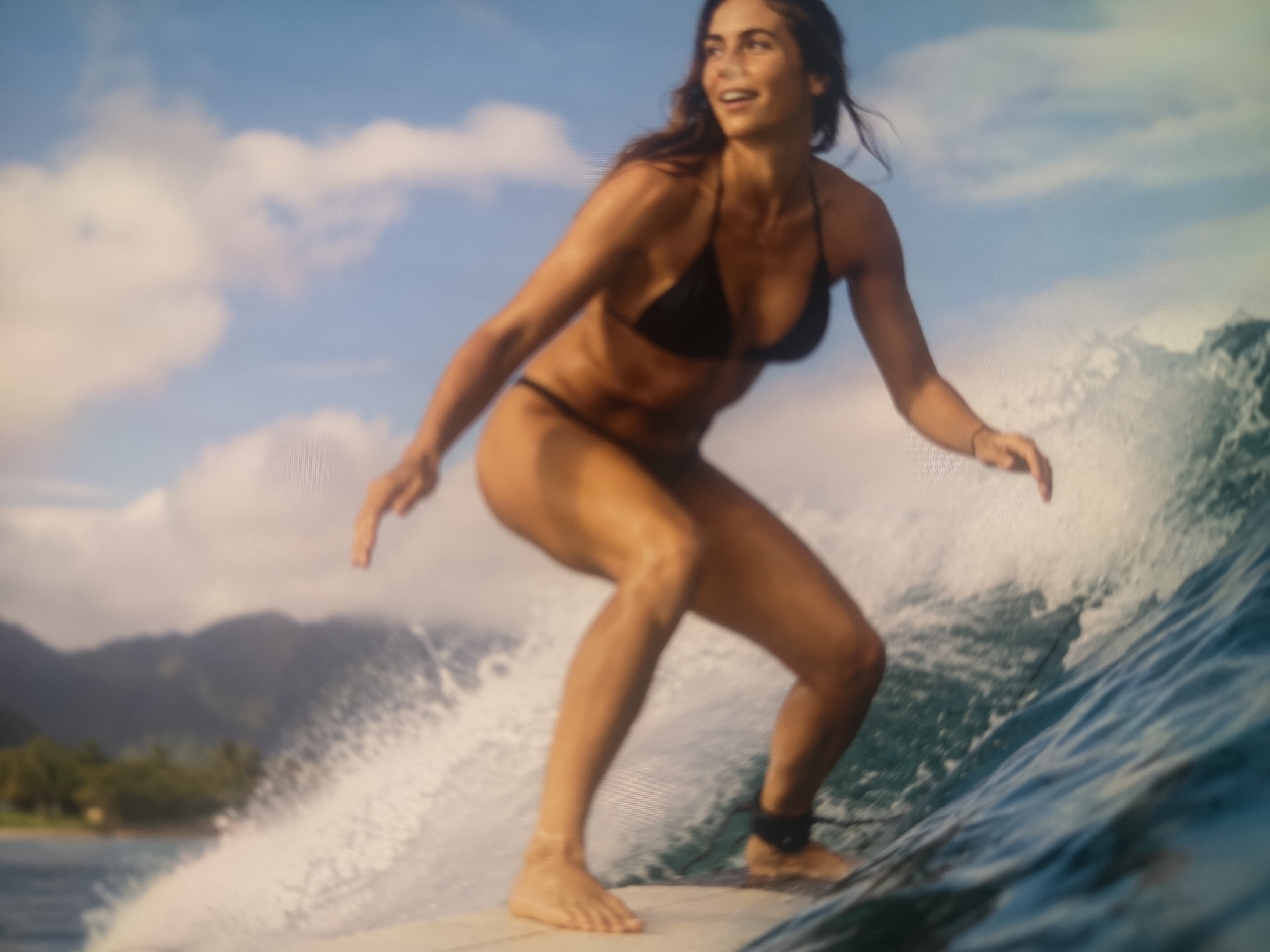 A woman is surfing on a wave. She is wearing a black bikini. The wave is breaking around her. In the background, there is a coastline with mountains. The location is likely a tropical area, possibly in the city of Unknown, Unknown.