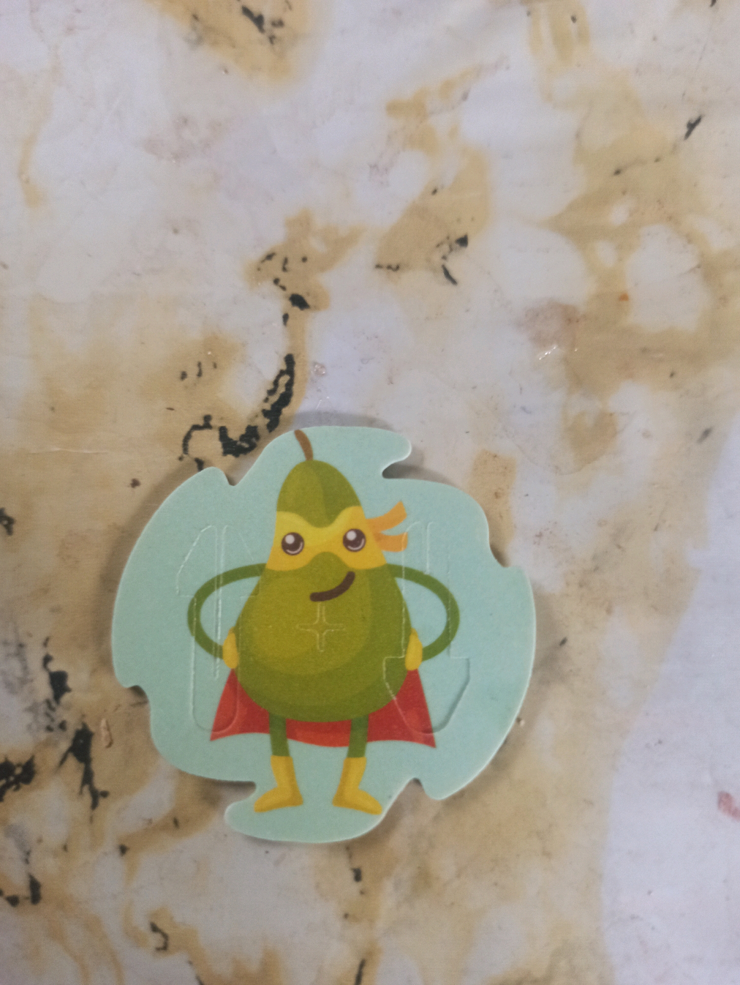 A cartoon avocado character, dressed as a superhero with a cape and mask, is positioned in the center of the image. The character has yellow boots and gloves, and is standing with its hands on its hips. The background is a mottled, light-colored surface with irregular brown and black markings. The image is taken from a slightly overhead angle. The location specified, Machraa El Ain, Morocco, is not visually discernible within the image.