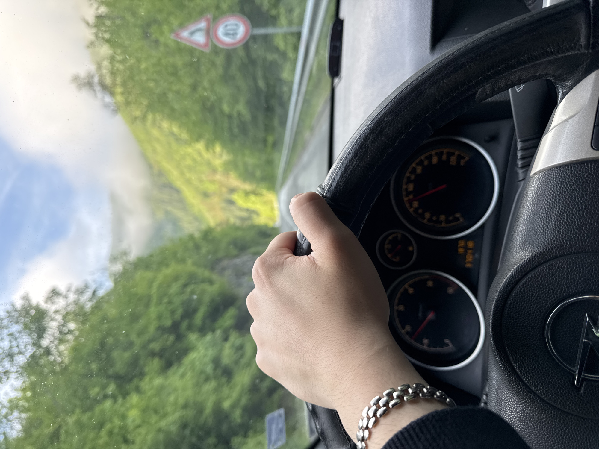 A person is driving a vehicle on a mountain road, with lush green vegetation visible through the windshield. A road sign indicating a speed limit of 40 is present. The driver's hand is on the steering wheel and a silver bracelet is on the wrist. The dashboard features a speedometer and other gauges. The logo of a car manufacturer is visible on the steering wheel. The location is in the city of Taran, Bulgaria.