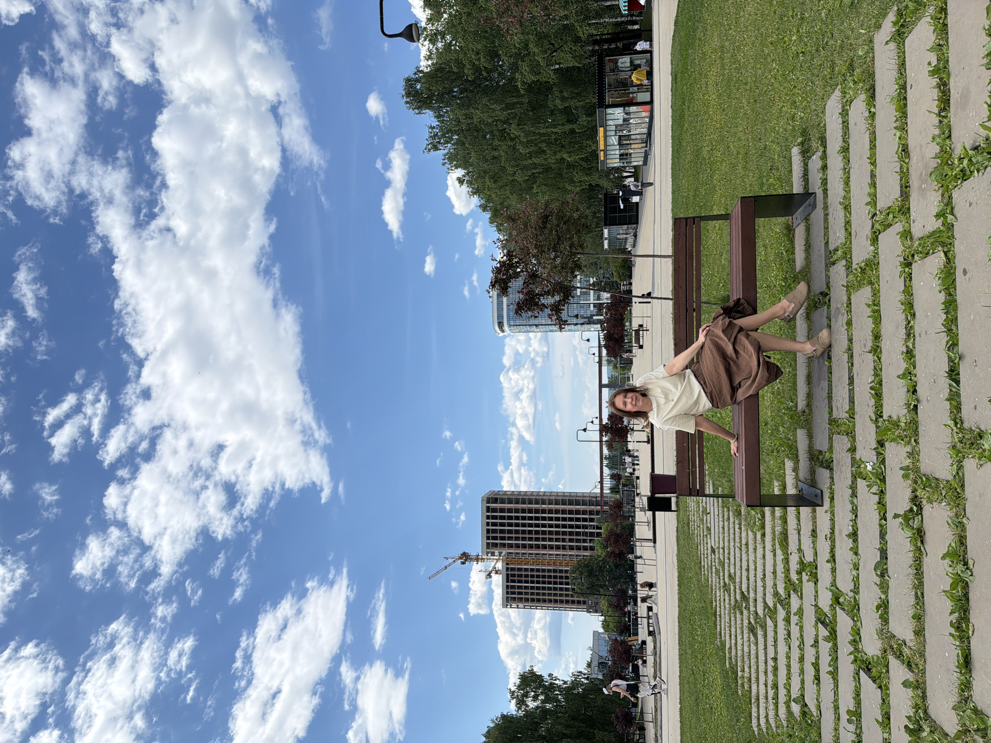 The image depicts a woman sitting on a park bench in a modern urban park. She is wearing a light-colored t-shirt and a brown skirt, with light brown slip-on shoes. The park features a pathway made of large concrete pavers interspersed with grass. In the background, there are trees, buildings under construction with cranes, and other people walking in the distance. The sky is blue with scattered white clouds. The location is identified as the "Golden Wave" Embankment Park in Izhevsk, Russia, which is a part of the new urban development along the Izh River.