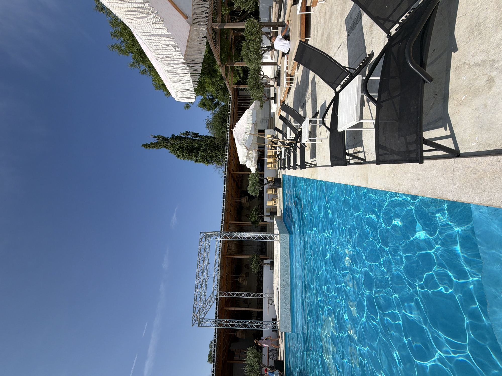 The image depicts a serene outdoor swimming pool area. In the foreground, a vibrant blue pool with rippling water is visible, reflecting the bright sunlight. Alongside the pool, black lounge chairs with small white side tables are neatly arranged on a light-colored paved surface. In the background, a wooden structure with a thatched roof serves as a covered seating area. A metal stage structure stands in front of this building, extending partly over the pool. A person in a pink dress and a straw hat walks near the stage, while another person in a red shirt is partially visible on the left. White umbrellas with decorative fringes provide shade over some of the lounge chairs. Green trees and olive trees are scattered around the perimeter of the area, adding to the natural ambiance. The sky is a clear, bright blue with a few wispy clouds. This scene appears to be at a resort or a similar leisure facility. The image doesn't provide explicit clues to confirm its location in Burgas, Bulgaria.