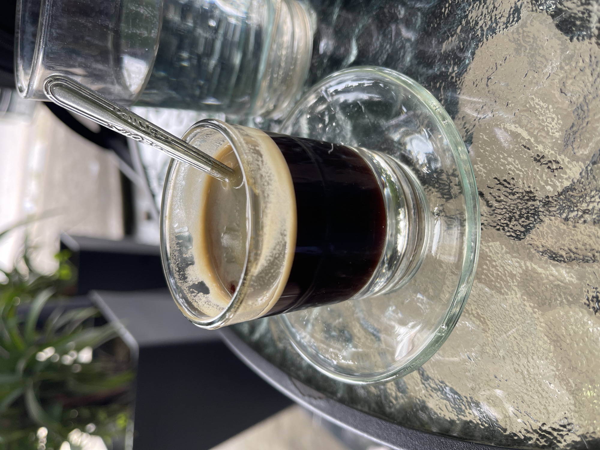 A close-up shot shows a small glass of espresso with a spoon in it, sitting on a glass saucer on a textured glass table. To the right of the espresso is a glass of water. In the background, out of focus, are two large black planters containing green plants. The setting appears to be outdoors, possibly at a cafe or restaurant, in Pantegajah, Indonesia.
