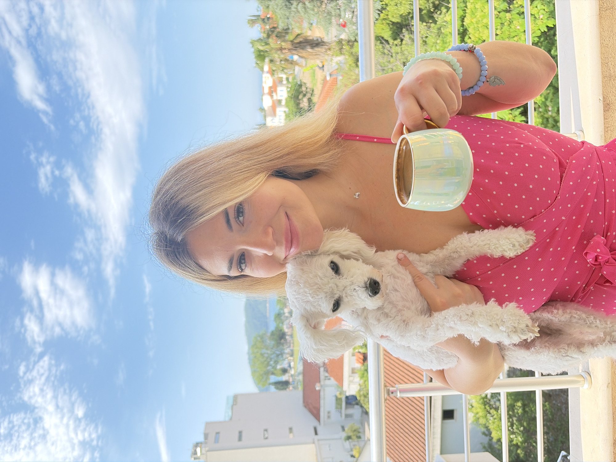 A woman with blonde hair is holding a white, fluffy dog while standing on a balcony. She is wearing a pink polka-dot dress and holding a light blue mug in her right hand, displaying several beaded bracelets on her wrist.
The background features a view of Budva, Montenegro, with residential buildings, trees, and a clear blue sky with scattered clouds visible. The perspective suggests the photo was taken at a higher elevation overlooking the town. A metal railing surrounds the balcony, further establishing the location.