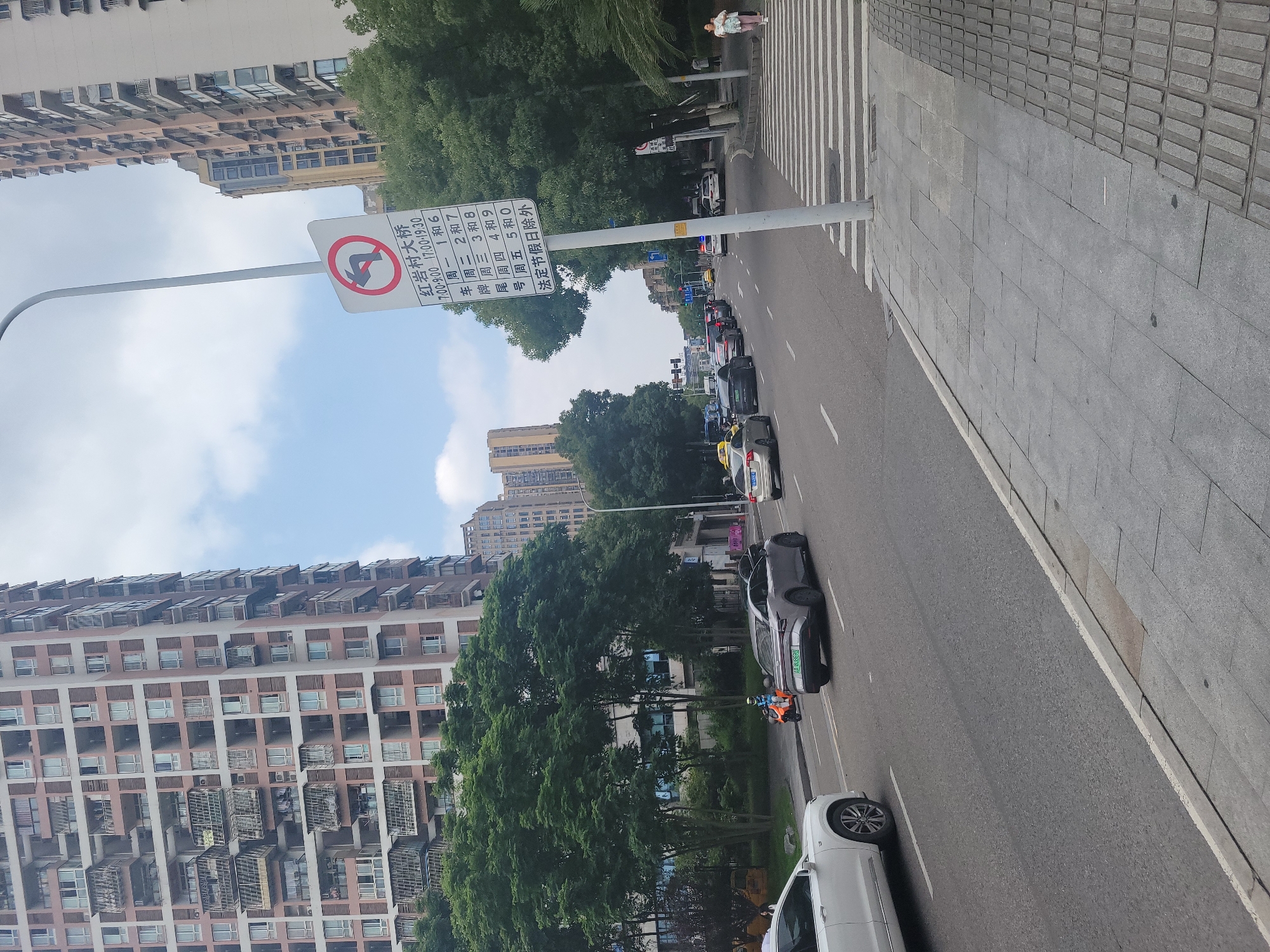 The image depicts a city street scene in Yuzhong District, China, on a bright, overcast day. On the left side of the image are tall, multi-story apartment buildings with numerous windows and balconies. Lush green trees line the street in front of the buildings. The street itself is paved with asphalt and marked with white lane lines. Several cars are driving on the road, including a white SUV on the left, a gray electric car with license plate "AA BL8828" in the middle, and other vehicles further down the road. A scooter with a rider wearing an orange jacket is visible on the left side of the road.
A prominent traffic sign on a pole is positioned in the middle right of the image. The sign has a red circle with a diagonal line indicating a prohibition, and an arrow curving to the left, suggesting a no left turn or a restricted turn. Below this symbol, there is text in Chinese characters, which appears to be traffic regulations and specific times, followed by "法定节假日除外" meaning "except for statutory holidays."
To the right of the road is a sidewalk paved with gray tiles, featuring tactile paving for the visually impaired. A pedestrian crossing with white stripes is visible on the road leading to the sidewalk. Further down the street on the right, another tall apartment building can be seen. A woman is walking on the sidewalk on the far right of the image, carrying a child. The sky is partly cloudy.