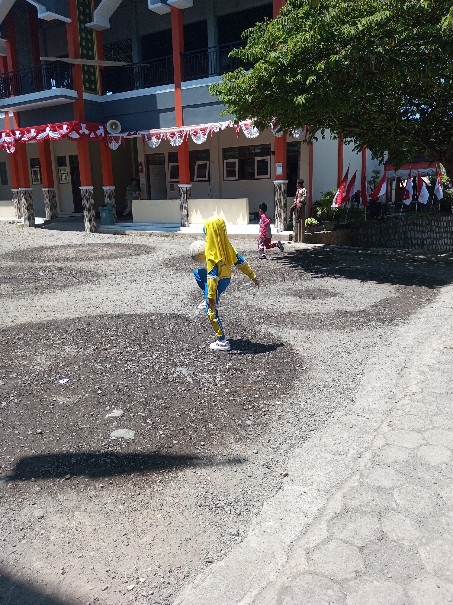 In Krasak, Indonesia, a young girl wearing a yellow hijab and blue and yellow sportswear is depicted playing with a volleyball in a schoolyard. The ground is covered in dirt and gravel, with a paved area to the right. In the background, a two-story building with orange pillars and white and red decorative banners suggests a school or public facility, possibly decorated for an event. Several red and white Indonesian flags are visible on poles near the building, reinforcing the national context. Two other children, one in a pink shirt and the other in a school uniform, are also present in the scene, with an adult in a uniform standing near them. The overall atmosphere is bright and sunny, indicating a daytime setting.