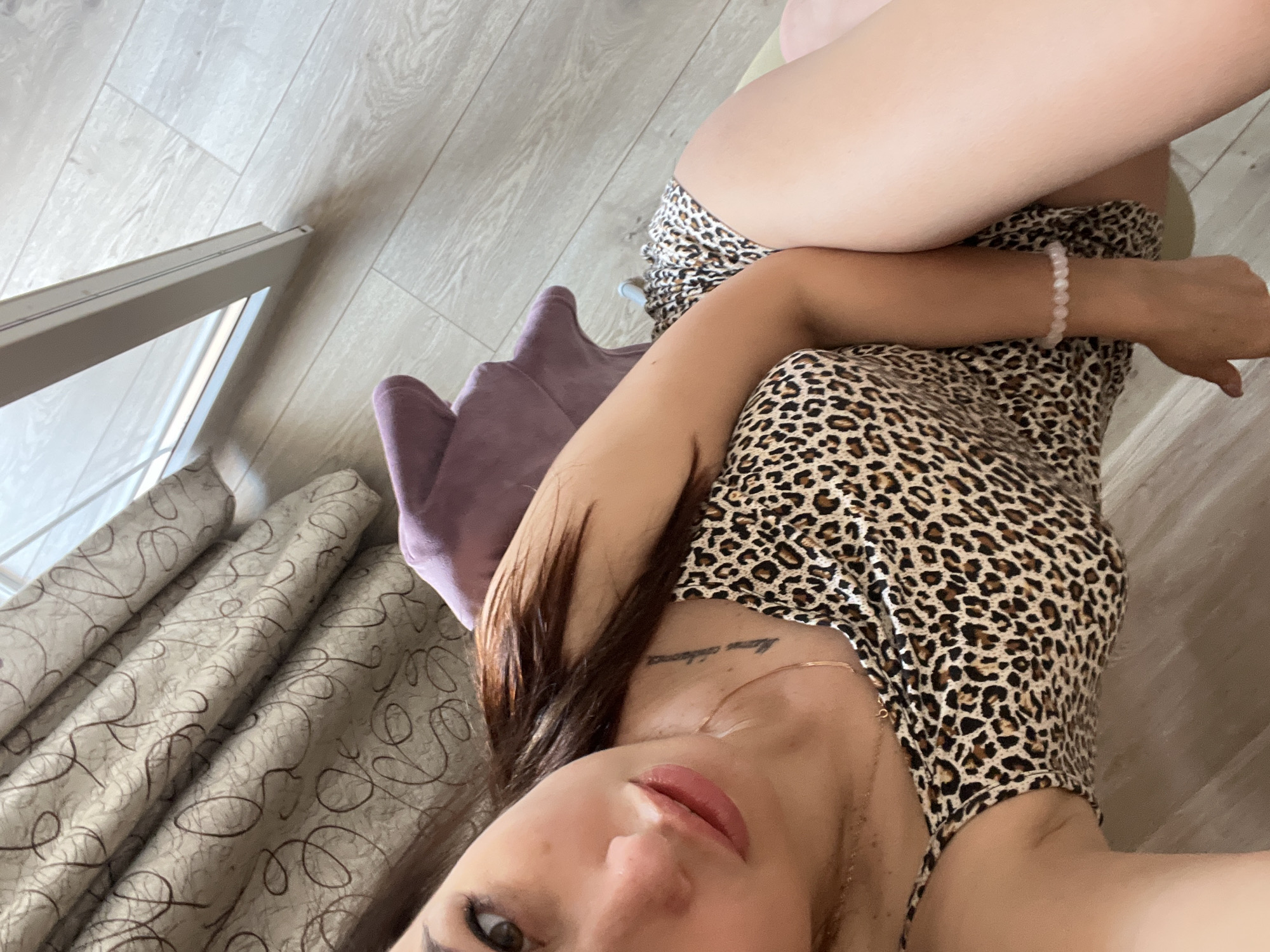 A high-angle, close-up shot captures a young woman with long, dark hair reclining on a purple chair. She is wearing a leopard-print camisole and matching shorts. Her left arm rests across her body, and she has a light pink beaded bracelet on her wrist. A delicate gold chain necklace is visible around her neck. A tattoo is present on her collarbone. The background reveals beige curtains with a dark, swirly pattern and light-colored wooden flooring. A portion of a window frame is visible on the right side of the image. The lighting appears to be natural, coming from the window.