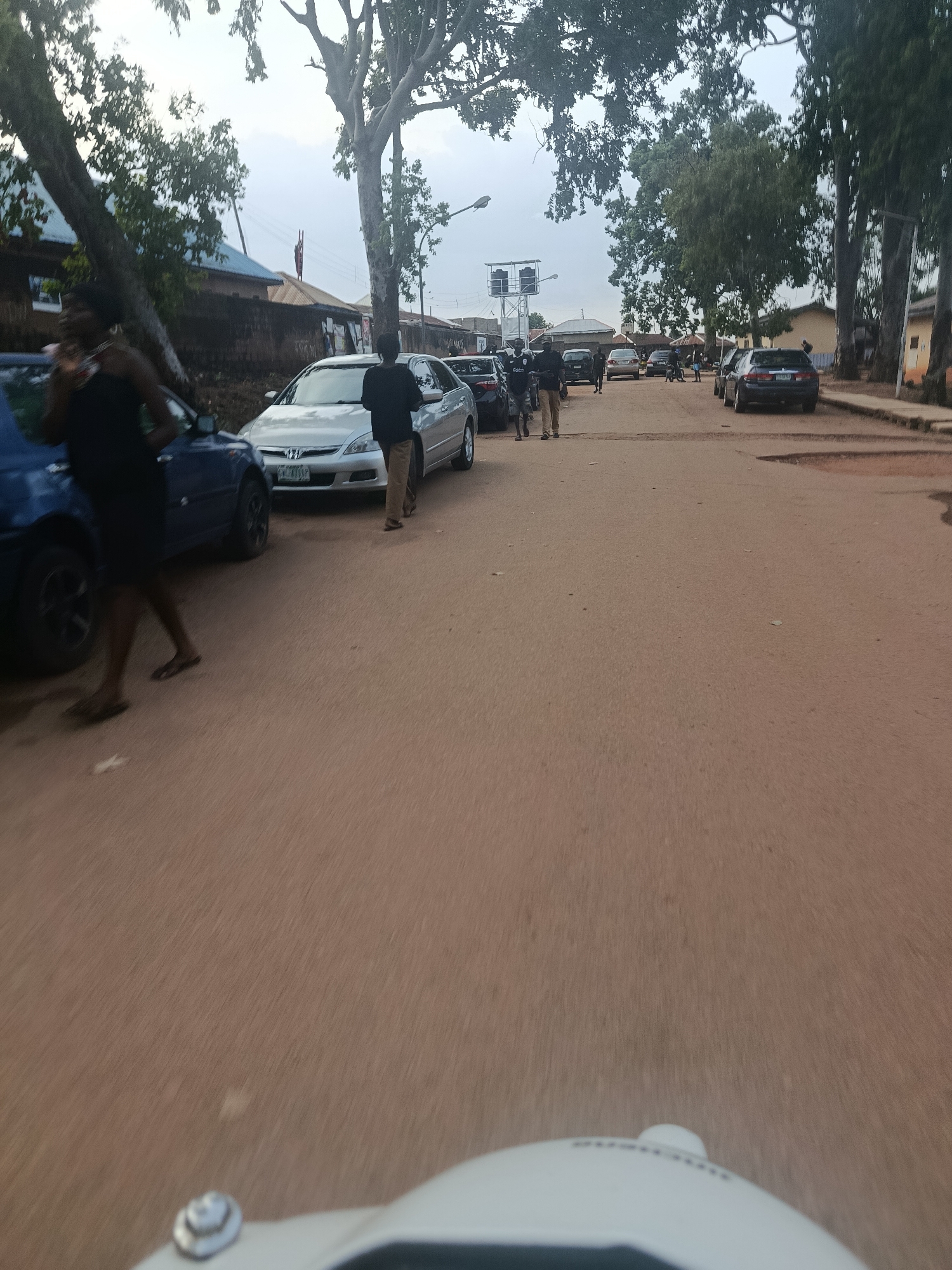 The image depicts a street scene in Rafin Par, Nigeria. The road is unpaved and appears to be made of reddish-brown earth. Several cars are parked along the side of the road, and some are in motion. A few people are visible, walking on the sidewalk and near the cars. Trees with large canopies line the street, providing shade. The sky is overcast, suggesting a cloudy day. The overall impression is of a busy, everyday street in a Nigerian town. The camera is at a low angle, possibly mounted on a vehicle, and the motion blur suggests the photo was taken while moving.