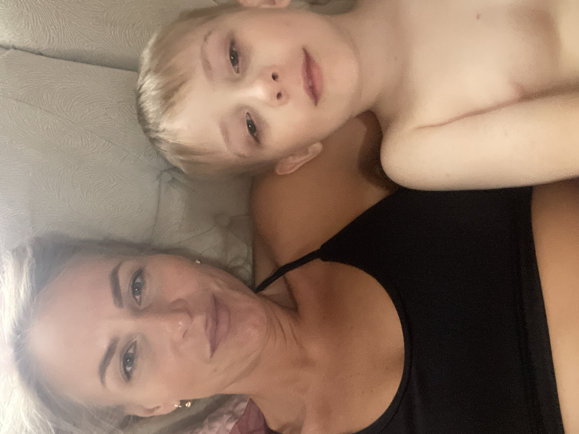A woman and a young child are lying on a light-colored surface, possibly a couch or bed. The woman is wearing a black tank top. The child is fair-skinned with blond hair and appears shirtless. They are positioned close to each other, likely in an embrace. No information suggests this image was taken in Baranovka, Russia.