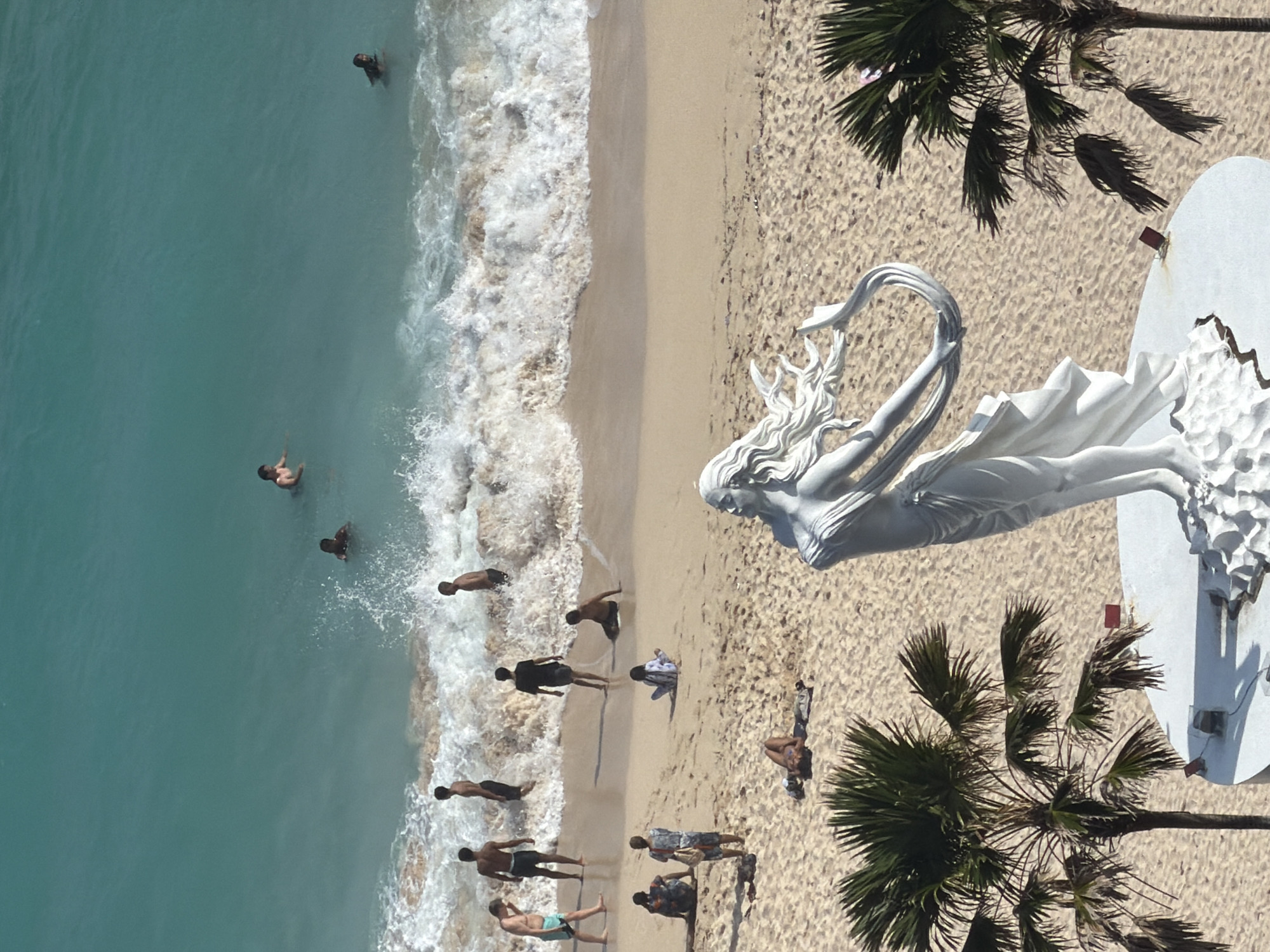 An aerial view captures Melasti Beach in Ungasan, Bali, Indonesia, under bright daylight. In the immediate foreground, a prominent white, neoclassical-style statue of a female figure with flowing hair and drapery stands on an elevated circular pedestal. Two palm trees partially frame the lower left and right corners of the pedestal.
The sandy beach, light in color, extends behind the statue towards the turquoise ocean. On the sand, a total of six individuals are observed. One person is lying prone near the center-left, facing away from the camera. Three individuals are seated; two are positioned close to each other, and one is seated with their back to the water. Two other individuals stand further back on the sand, closer to the water's edge.
Breaking waves with white foam mark the transition from the sand to the ocean. Within the shallow water, five individuals are seen standing or wading, interacting with the incoming waves. Further out in the calmer, deeper turquoise water, two additional individuals are swimming or floating. The overall scene depicts a recreational beach environment with multiple people engaged in various leisure activities.