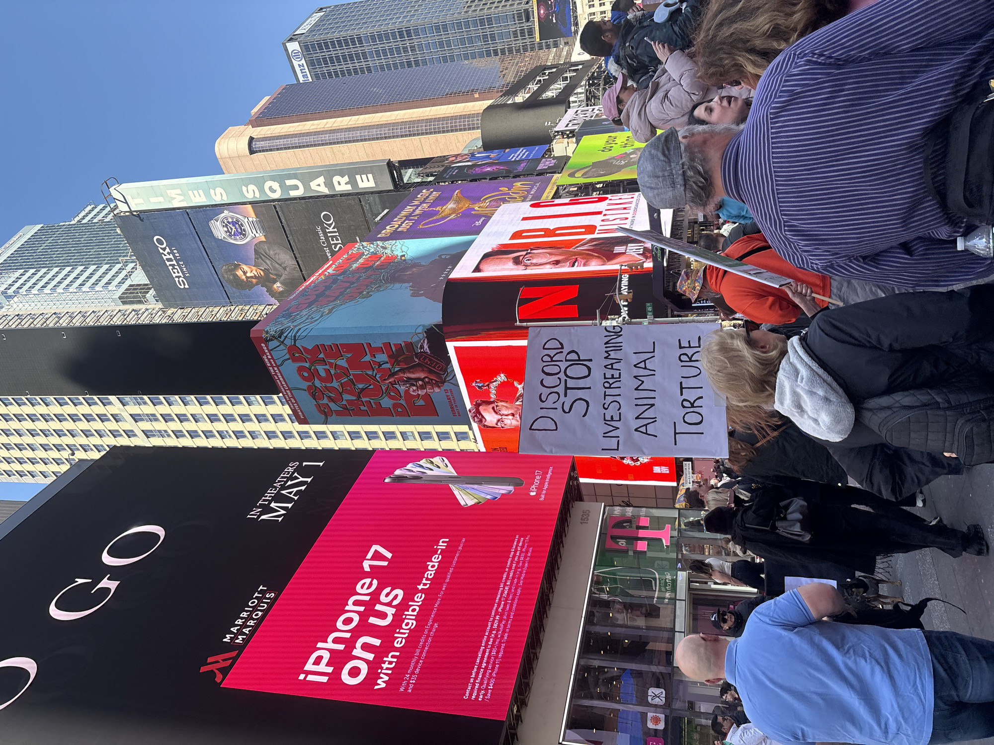 The image captures a bustling scene in Times Square, New York City, USA. People are gathered, some of whom are holding a sign that reads "DISCORD STOP LIVESTREAMING ANIMAL TORTURE," suggesting a protest or demonstration. The environment is characterized by numerous large, bright advertising billboards displaying various brands, including "iPhone 17" at Marriott Marquis, Seiko, and advertisements for movies and Broadway shows. The architecture consists of tall skyscrapers in the background, and the overall atmosphere is crowded and vibrant.