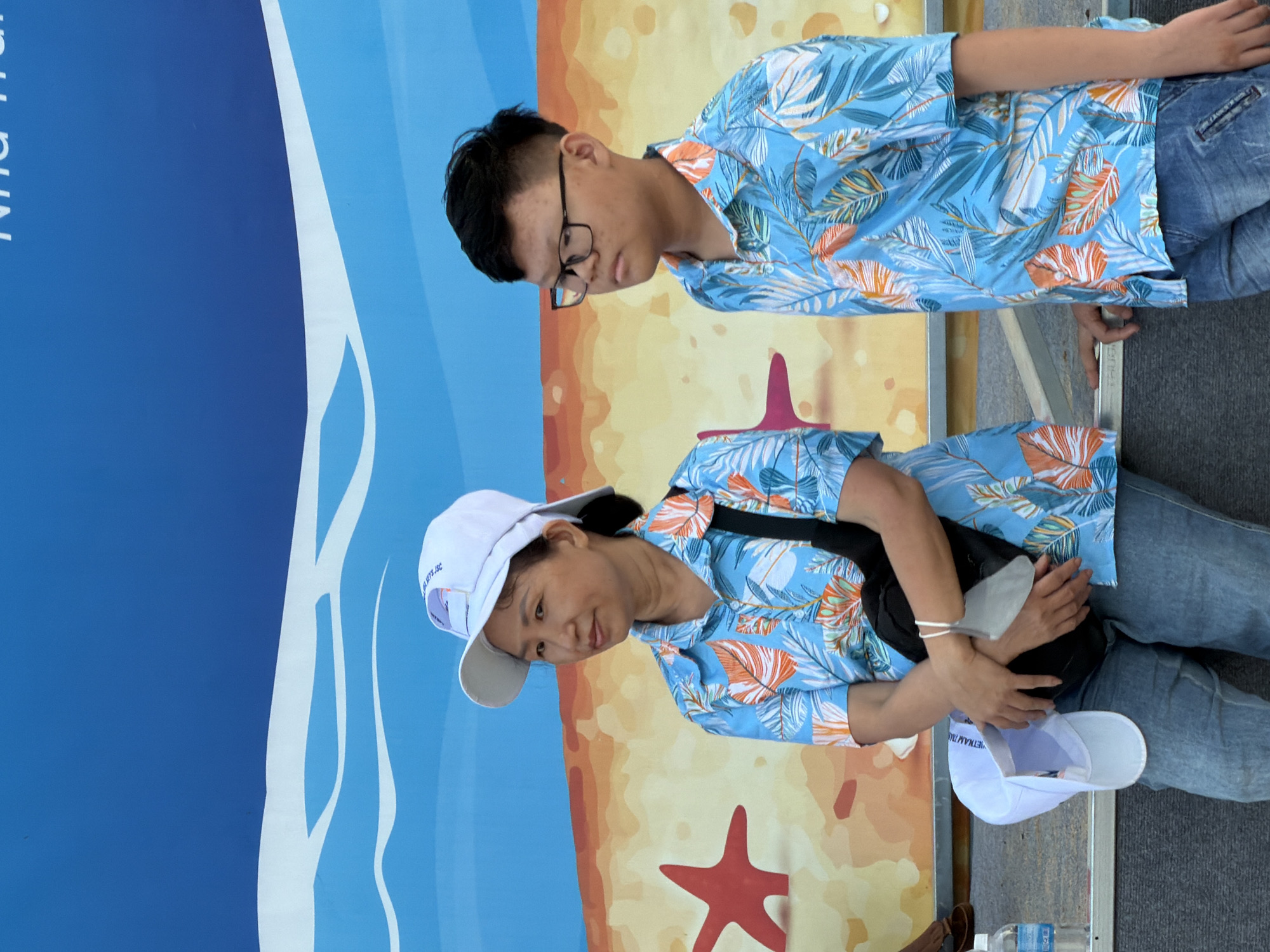 A woman wearing a white baseball cap and a blue floral shirt stands with her arms crossed, holding a white face mask and a black bag across her chest. To her right, a boy wearing glasses and a matching blue floral shirt looks to the right. They are standing in front of a blue backdrop with a cartoonish depiction of waves and a beach scene with starfish and sand. The scene appears to be outdoors, possibly at an event or tourist location in Nha Trang, Vietnam, given the text visible on the backdrop.
