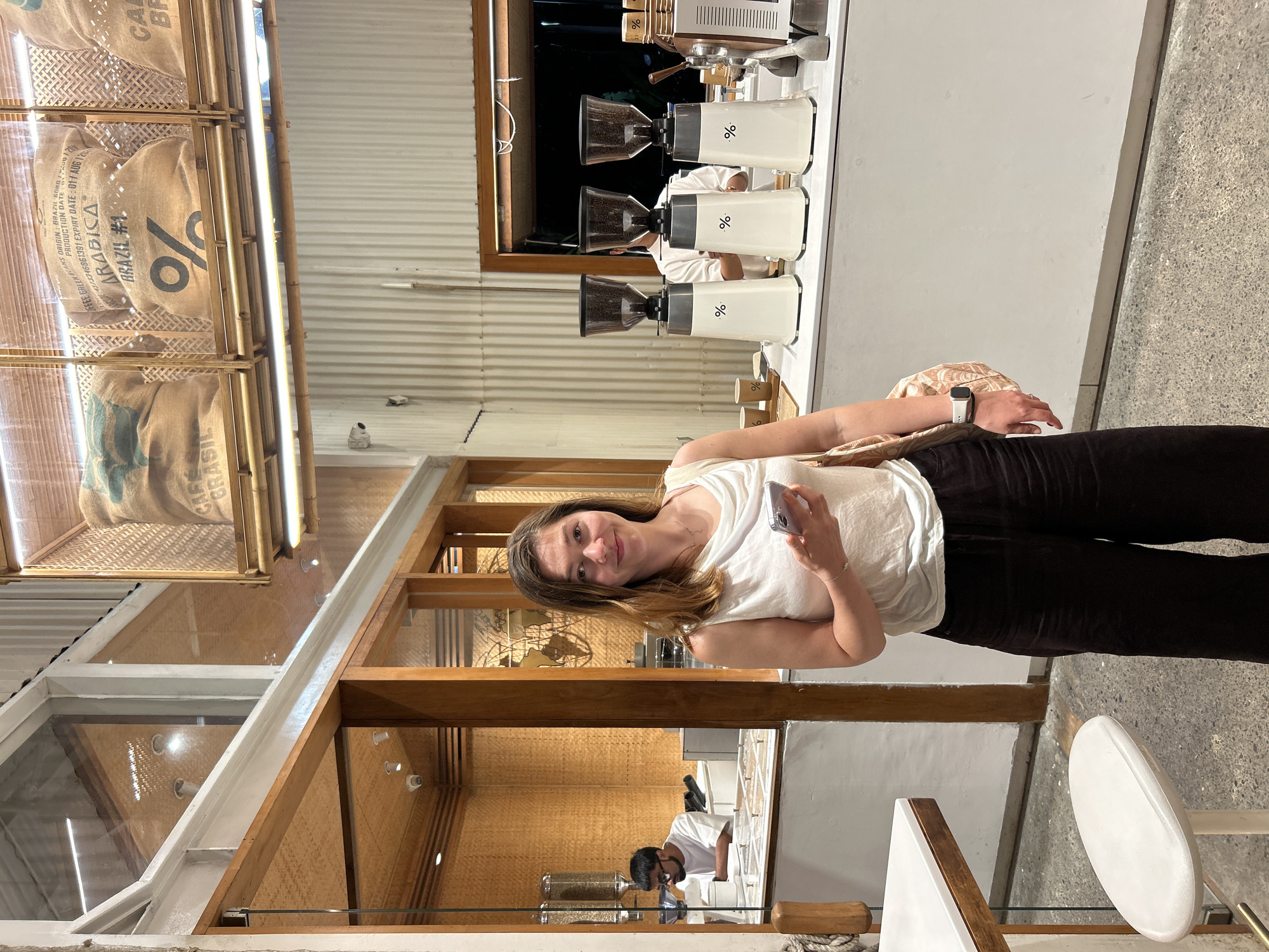 A woman with shoulder-length brown hair, wearing a white sleeveless top and dark wide-leg pants, stands in a coffee shop, holding a smartphone in her right hand and a patterned tote bag over her left shoulder. She is looking at the camera with a slight smile. Behind her, a counter displays three white coffee grinders with the percentage symbol logo. Further back, a man in a white t-shirt is visible behind the counter, wearing glasses. The coffee shop has a rustic interior with bamboo accents on the walls and ceiling, and burlap sacks labeled "ARABICA BRAZIL" are displayed on shelves above. The overall scene suggests a modern, artisanal coffee shop. The image appears to have been taken in Kuta Selatan, Indonesia, given the visual style and common aesthetics of establishments in that region.