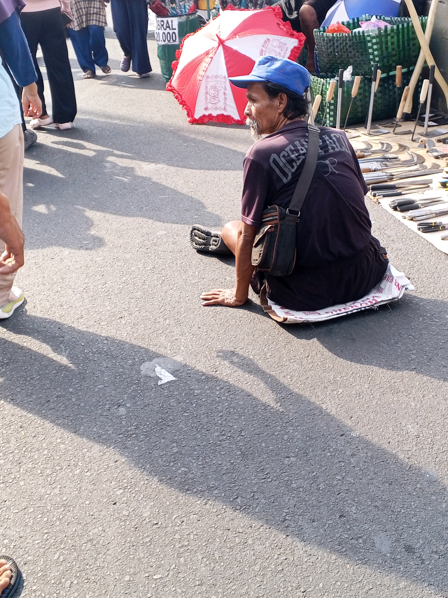 The image captures an outdoor scene on a paved surface, likely a street or market area in Serang, Indonesia. In the foreground, a man wearing a blue baseball cap and a dark t-shirt is seated on a white mat. He has a bag slung across his chest. His attire suggests casual wear. Behind him, there is a display of various tools, including what appear to be knives and other sharp implements, laid out on white fabric. Several green woven baskets are visible in the background, along with a red and white umbrella. Other people are present in the scene, mostly their legs and lower bodies are visible, indicating they are standing and walking. Some individuals are wearing casual clothing, including pants and footwear. The ground is asphalt, and there are distinct shadows cast by the people and objects, suggesting it is a sunny day. The overall impression is that of a market stall or vendor setup, possibly selling tools or other goods.