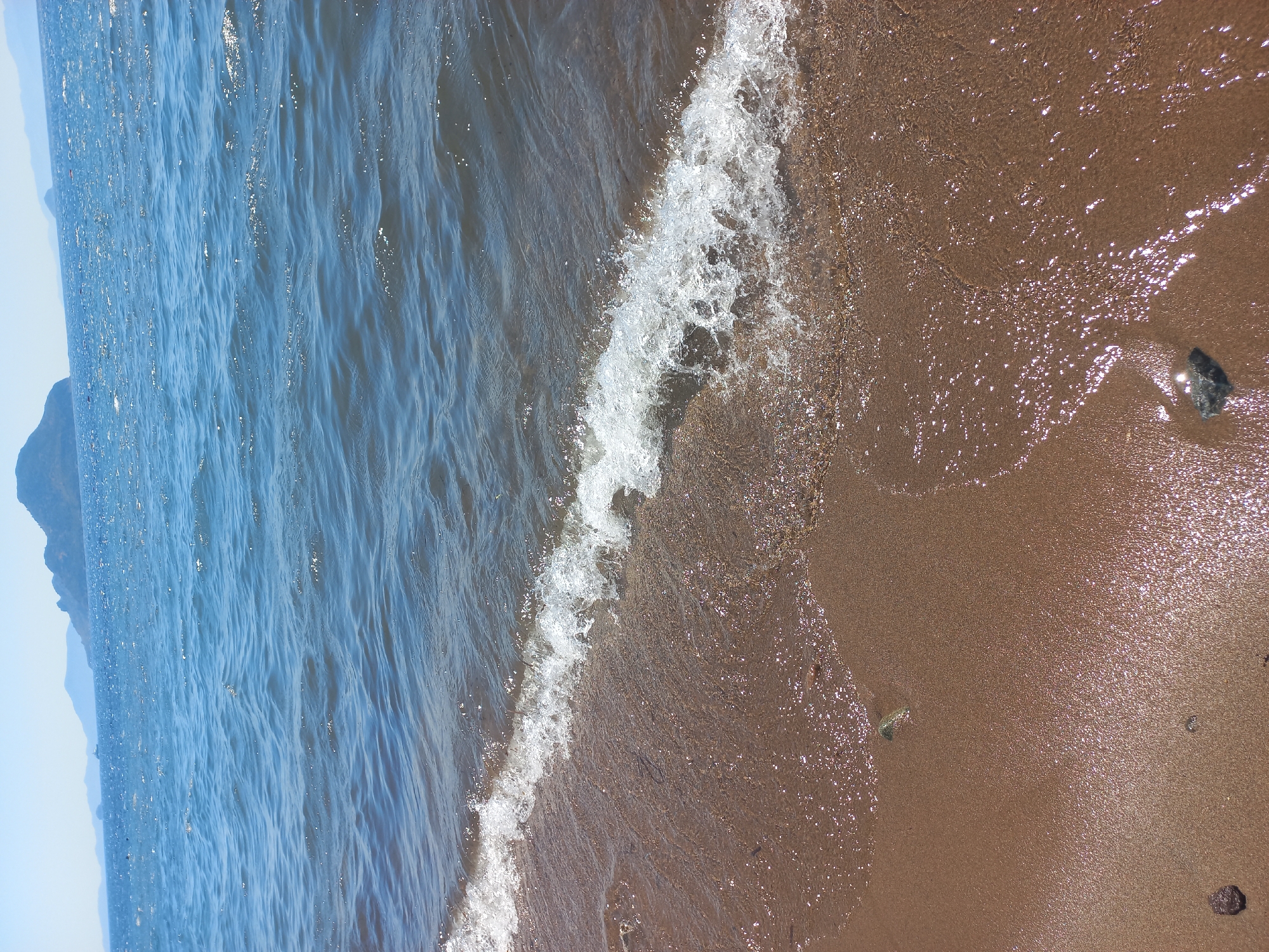 A photograph captures a close-up view of a shoreline in Bodrum, Turkey, under a bright, clear sky. The foreground is dominated by wet, sandy beach with scattered small pebbles. A gentle wave with white foam is breaking onto the shore, its water creating shimmering reflections on the sand. In the background, the Aegean Sea stretches out, its surface rippled and reflecting sunlight. Further in the distance, a chain of mountains and islands forms the horizon. There are no people visible in the image.