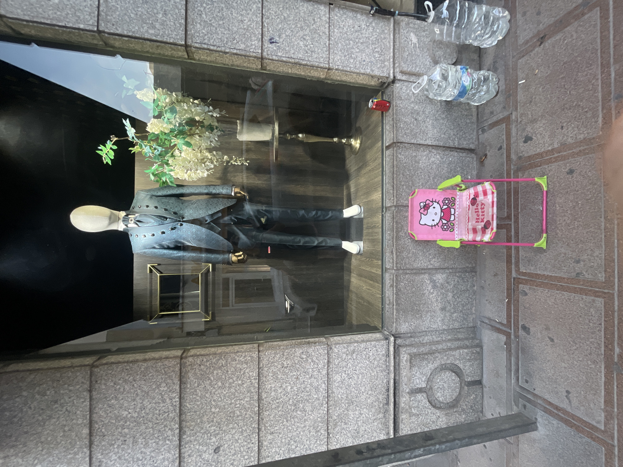 The image shows a storefront window display. Inside the window, a mannequin is dressed in a suit with a black tie and white shirt. White flowers are arranged behind the mannequin. To the right of the window, a pink toy stroller with a Hello Kitty design is visible, along with two plastic water bottles. The storefront is made of stone blocks, and the ground is paved. A metal railing is in the foreground. The scene appears to be set during the day, with natural light reflecting on the glass. There are no visible signs of weather conditions. The setting suggests a commercial area, likely a clothing store or boutique. The image is oriented sideways.