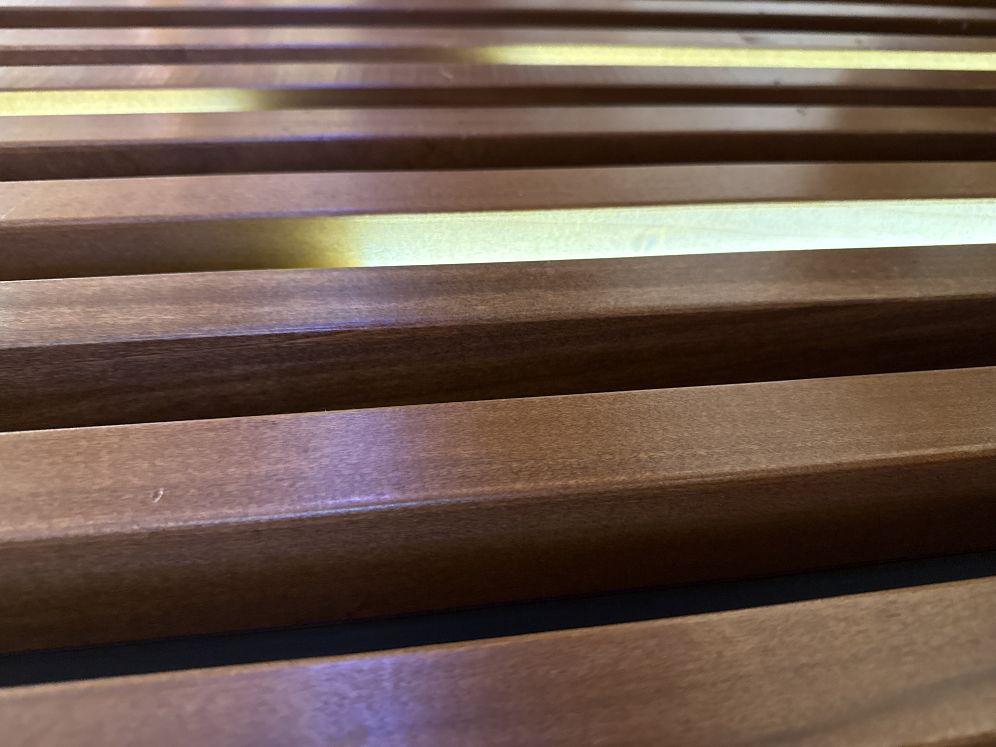 The image shows a close-up, angled view of parallel, horizontal wooden slats with light shining through the gaps between them. The wood is a medium brown color. The perspective suggests the viewer is looking upwards at the slats. The scene appears to be an architectural element, potentially a wall covering or screen. There is no evidence in the image to suggest the location in Hollywood Seminole Reservation, United States of America. The image contains no people, subjects, or actions.