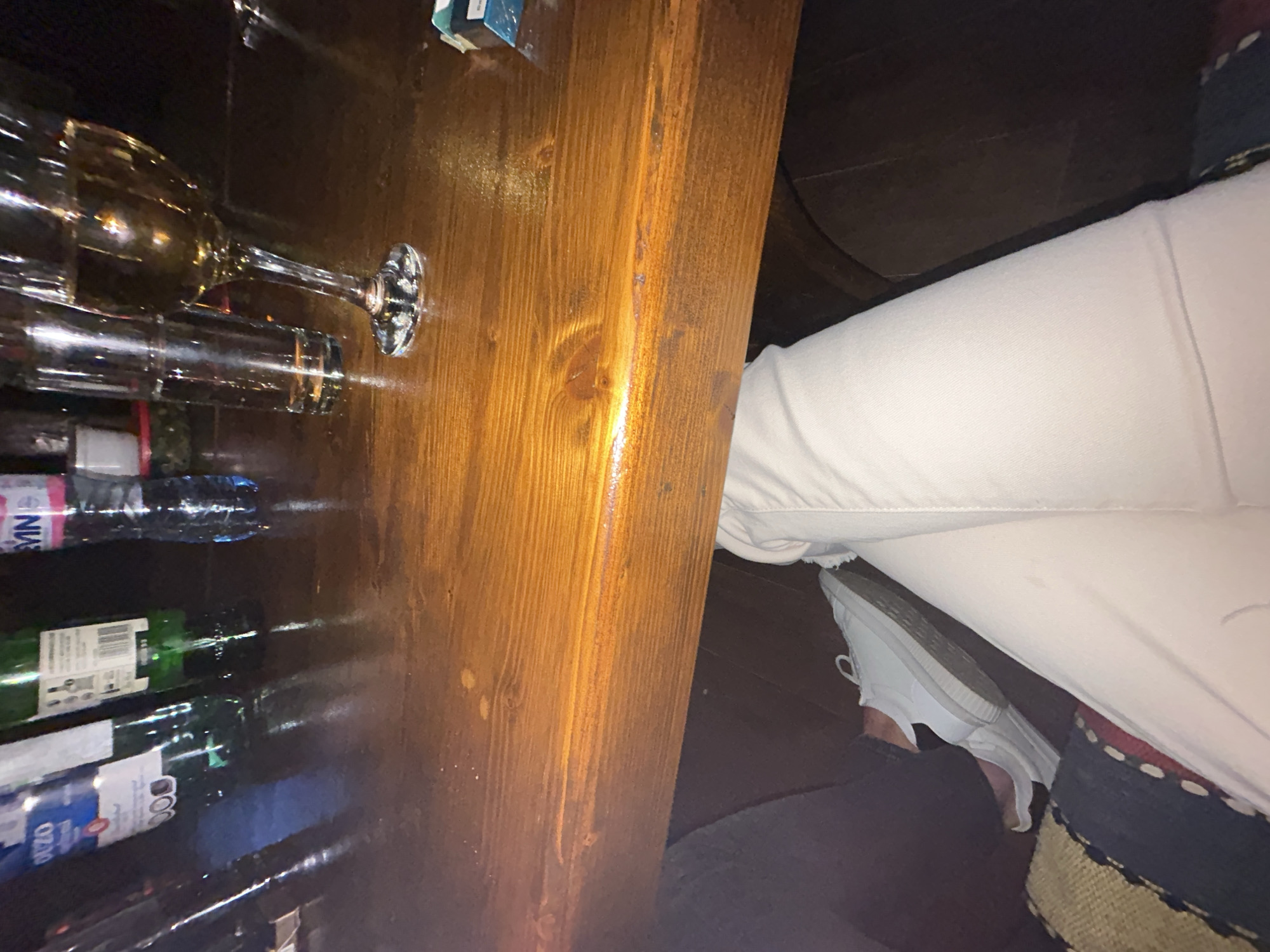 The image depicts a partially visible scene appearing to be within a bar or restaurant. A wooden bar top dominates the left side of the frame, cluttered with bottles of alcohol and a wine glass. A portion of a person's lower body is visible on the right, including white pants and a white sneaker. The person is seated at the bar. The setting is implied to be in Mirovyane, Bulgaria.