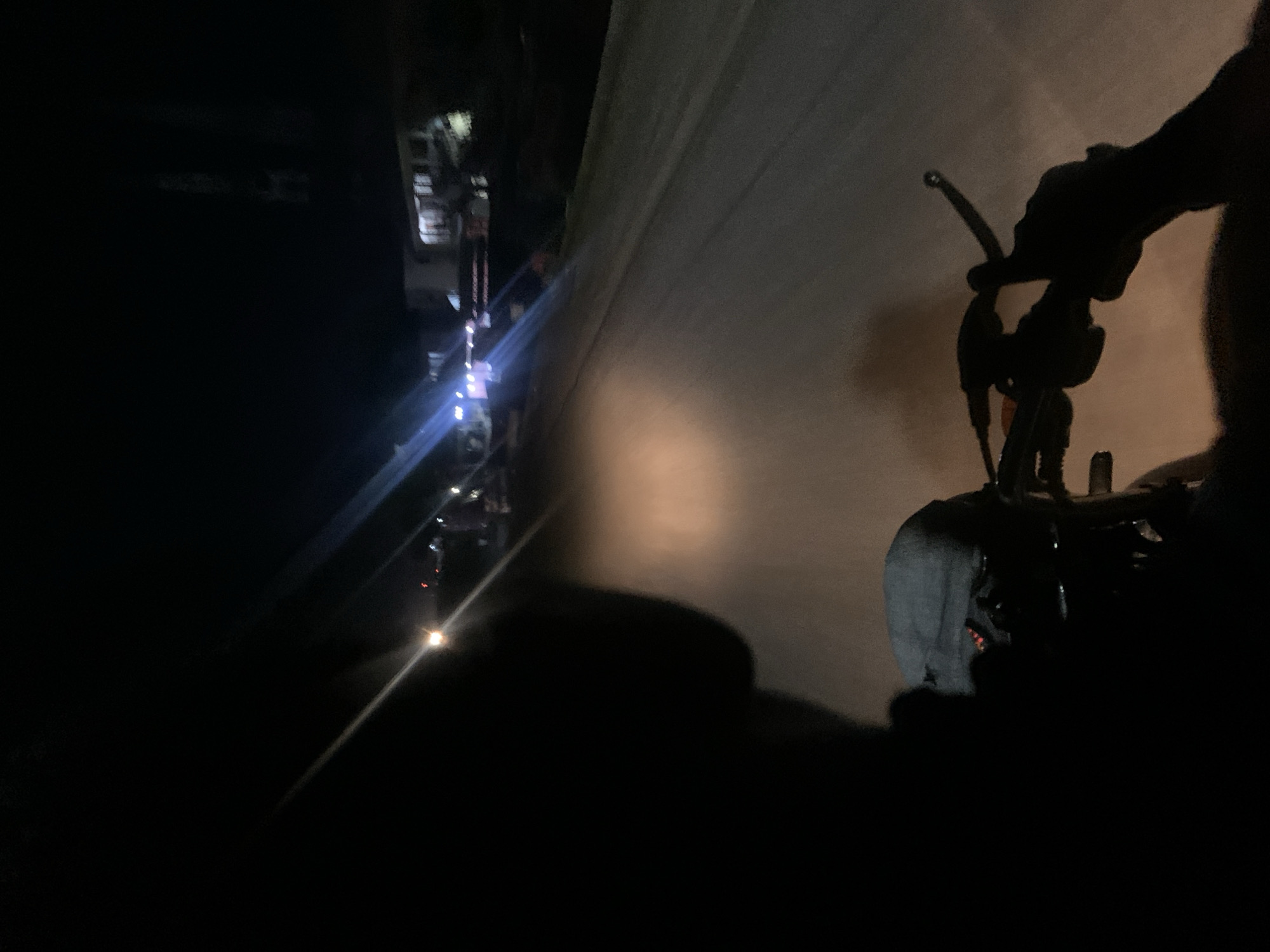 A person rides a bicycle at night on a road in Adankolo, Nigeria. The road is lit by the bicycle's headlight, which illuminates a section of the road ahead. Street lights in the distance suggest a built-up area. The rider is wearing a gray hoodie.