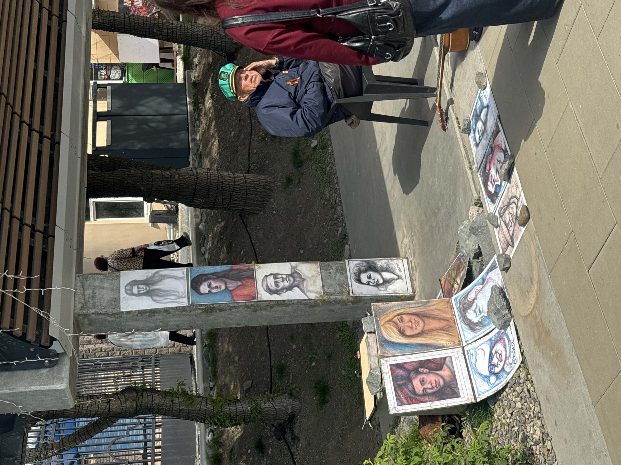 The image depicts an outdoor art scene in Vladivostok, Russia. An elderly man wearing a green sailor's cap and a dark jacket is seated in a black plastic chair next to a sidewalk. He appears to be interacting with a passerby, whose upper body is visible, wearing a maroon jacket and carrying a black purse.
Displayed along the sidewalk and on an adjacent low wall are numerous framed portraits, likely the artist's work. The portraits feature various subjects, predominantly headshots and faces. Some are propped up by small rocks to prevent them from falling over.
A guitar is leaning against the chair, indicating the artist might also be a musician. The scene is set against a backdrop of trees, a building with wooden slats, a window, and a metal fence, all indicative of an urban environment. The sidewalk appears to be next to a small patch of gravel and some greenery.