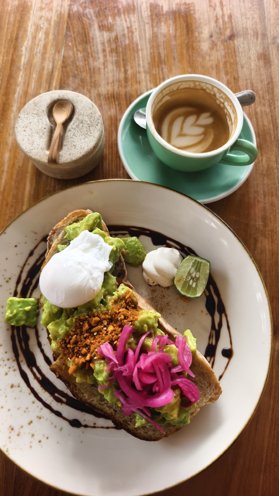 This overhead shot captures a vibrant breakfast or brunch setting on a warm, light brown wooden table. The main subject is a white plate featuring a slice of avocado toast, richly topped with mashed green avocado. A perfectly poached egg, with its smooth white surface, sits prominently on the avocado. The toast is further garnished with a sprinkle of reddish-brown, textured crumble (likely nuts or seeds) and a generous portion of bright pink pickled red onions, adding a pop of color. Dark, glossy sauce, possibly a balsamic reduction, is artistically swirled on the white plate beneath the toast. Accompanying the toast are a small dollop of white cream or sauce and a fresh lime wedge.
To the upper right, a light green ceramic cup filled with coffee, displaying intricate latte art in a leaf pattern, rests on a matching saucer with a silver spoon. In the upper left, a small, textured stone container with a tiny wooden spoon tucked into its opening suggests condiments like salt, sugar, or spices. The natural lighting indicates a bright daytime setting, consistent with a morning or early afternoon meal. The arrangement and presentation suggest a pleasant dining experience, likely at a cafe or restaurant in Kuta, Indonesia. No visible text is present in the image.
