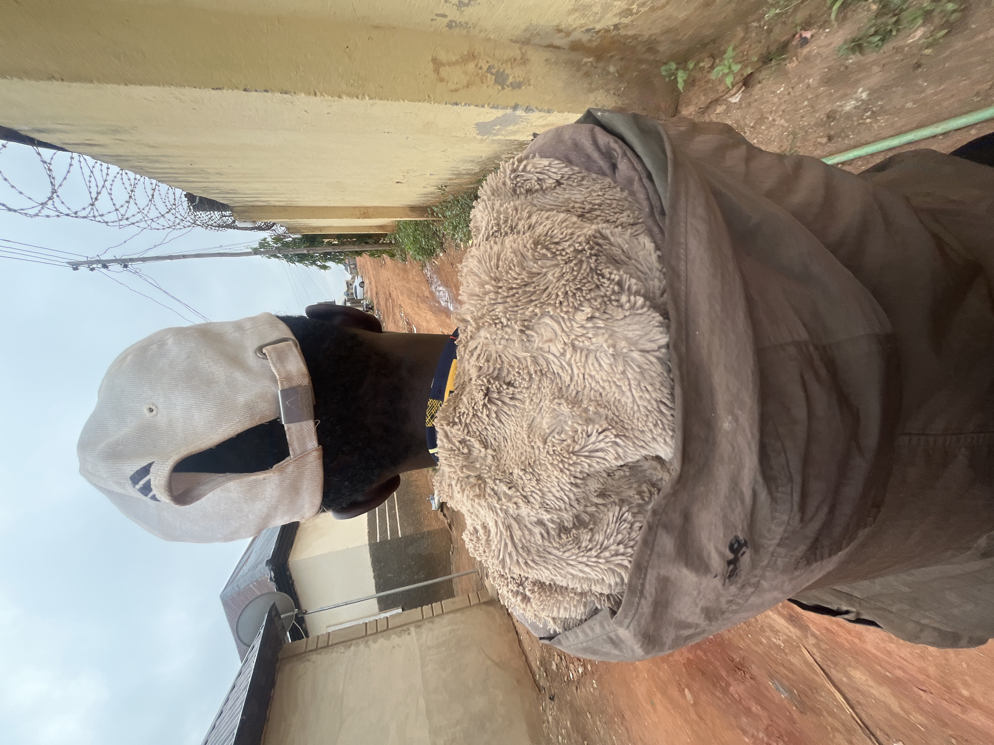 A person wearing a light-colored baseball cap and a brown jacket with a furry collar is standing in an alleyway in Galadimawa, Nigeria. The alley is lined with buildings on both sides. The ground appears to be muddy. Barbed wire is visible along the top of a wall.