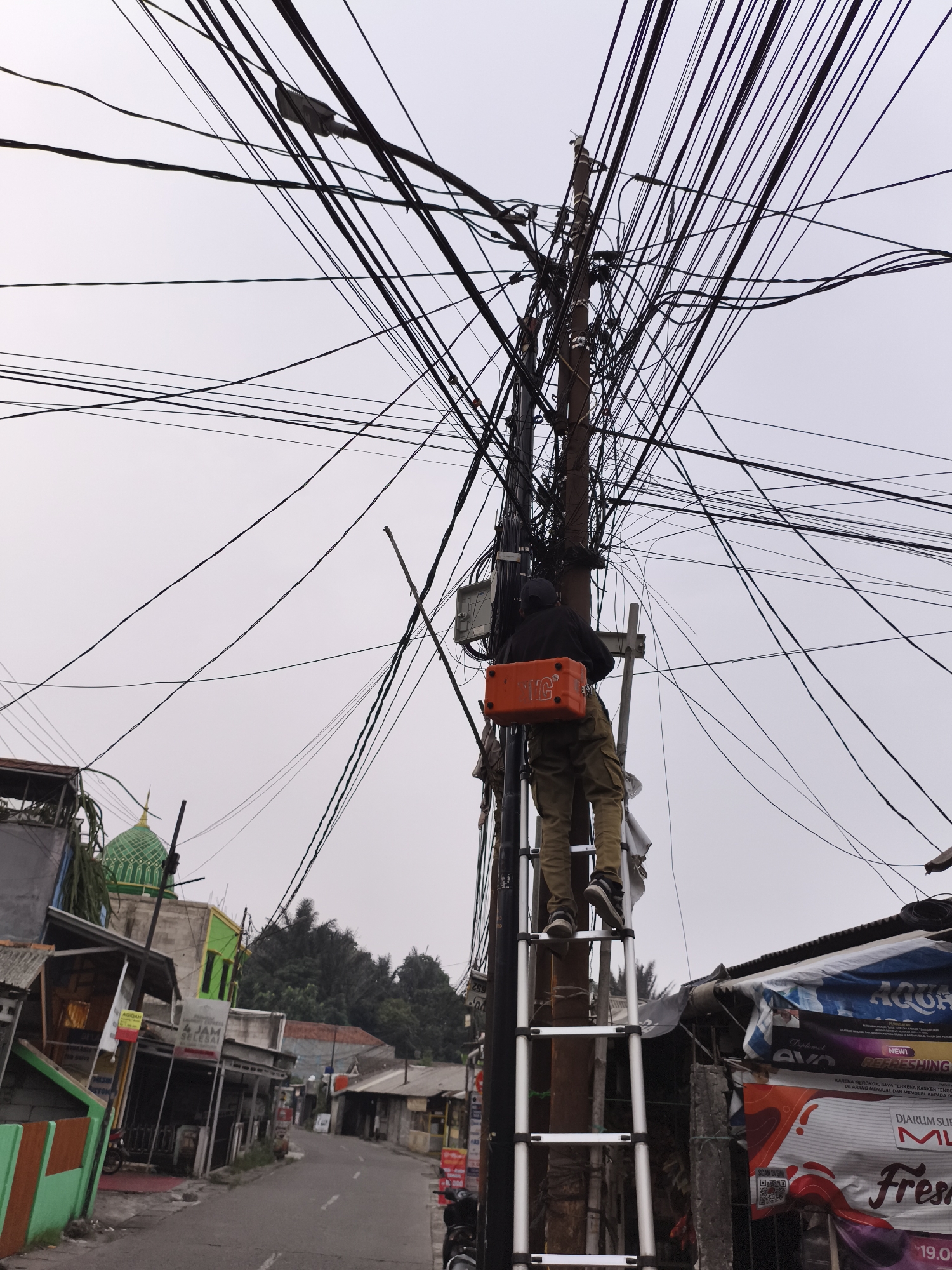 The image depicts a telephone repairman standing on a silver ladder while working on a utility pole densely packed with numerous crisscrossing wires in Depok, Indonesia. The repairman is wearing a black jacket, khaki pants, and dark sneakers. He has an orange toolbox strapped to his back. The pole is situated on a street lined with buildings on either side. On the left side of the image, a green-domed structure is visible above one of the buildings. A section of the street is visible between the buildings, and a motorcycle is parked in the lower portion of the image. On the right side of the image, a marketing sign is visible behind the utility pole that contains the words: "NEW! REFRESHING", "SCAN OS", "DJARUM SUPER", and "FRESH". The sky is overcast, and the overall lighting suggests it is either early morning or late afternoon.
