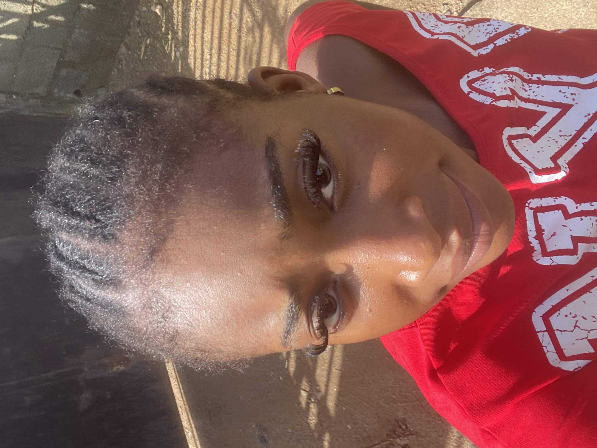 In Abuja, Nigeria, a young Black woman with braided hair is captured in a close-up, high-angle shot. She is wearing a bright red tank top with white lettering, partially obscured. Her face, illuminated by sunlight, shows a hint of a smile. Her eyes are adorned with dramatic, long false eyelashes, and she wears a small gold hoop earring in her visible ear. The background is softly blurred, suggesting an outdoor setting with elements of concrete and possibly fencing. The ground appears to be dirt or gravel, with shadows cast from an unseen source.