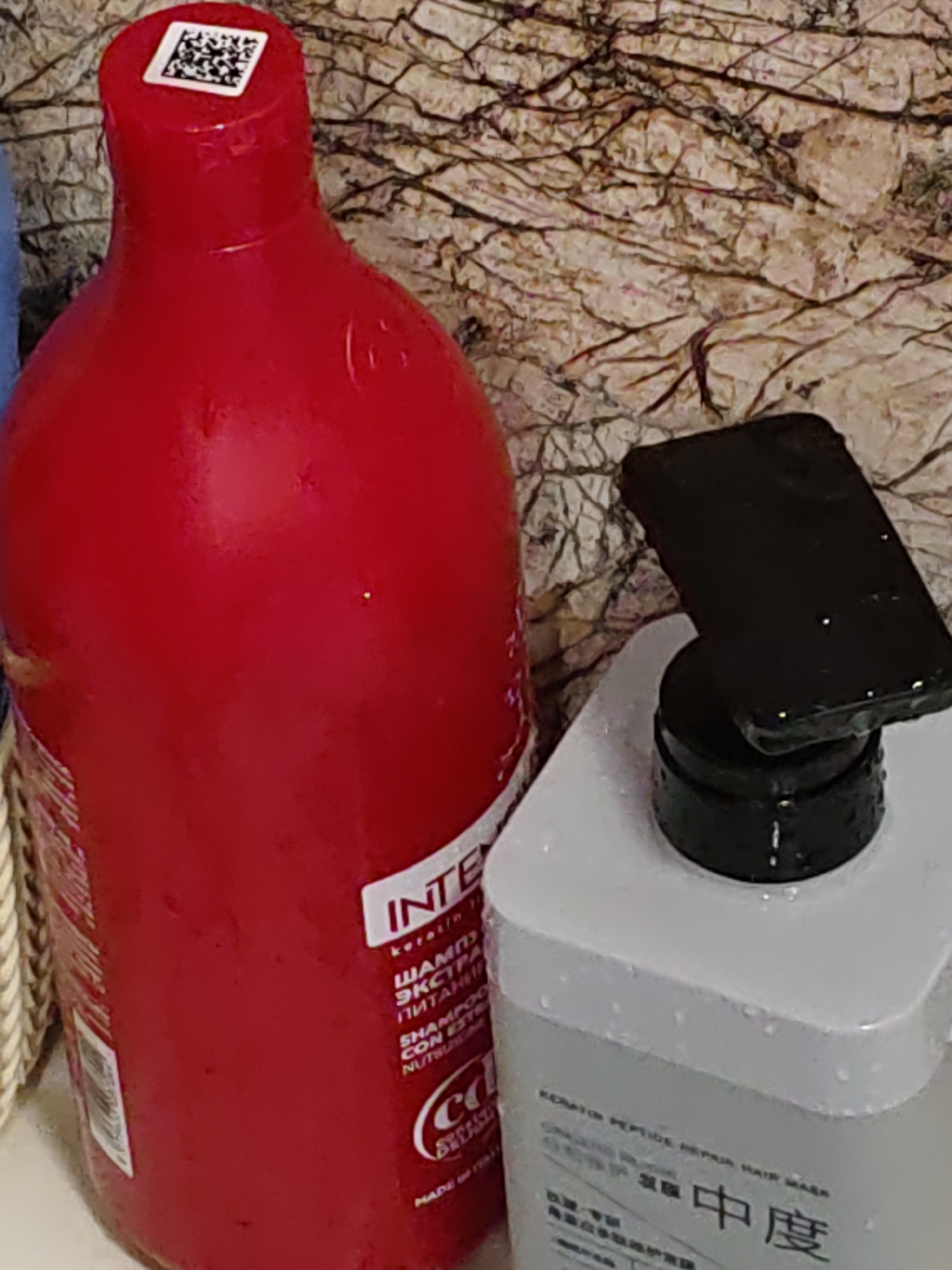 The image shows two bottles on a textured surface, likely a countertop. The bottle on the left is a large, red plastic bottle with a white label that has text in both Cyrillic and Latin alphabets. A QR code is visible on the red cap. The bottle on the right is a white plastic bottle with a black pump dispenser. A dark-colored object, possibly a phone case, rests on the dispenser. The scene is illuminated by artificial light, casting shadows and highlighting water droplets on the surfaces of the objects. The background appears to be a textured wall with a pattern resembling cracked stone. There is no visible indication of the city of Rostov-on-Don, Russia, within the image itself.