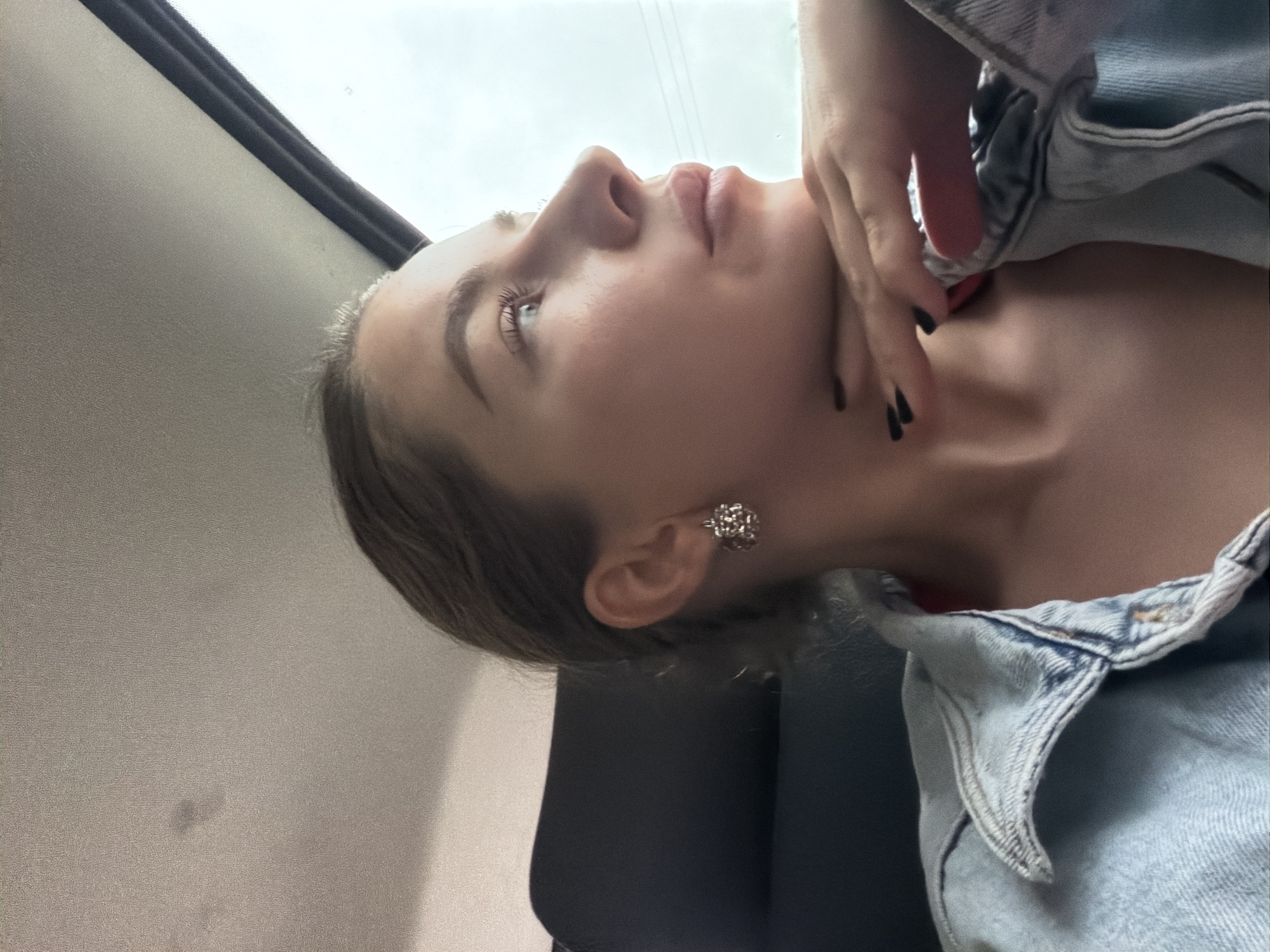 A young woman is shown from the chest up, in profile, looking out of a car window. She has fair skin, dark eyebrows, and dark eyelashes. Her lips are slightly parted, and she has her right hand resting on her chin. She is wearing a light blue denim jacket over a red top. Her fingernails are painted black. She has a silver earring in her right ear. The interior of the car is visible behind her, with a dark seat and the grey headliner of the car. The scene appears to be a bright, overcast day. The location is not identifiable as Gornji Vakuf-Uskoplje.