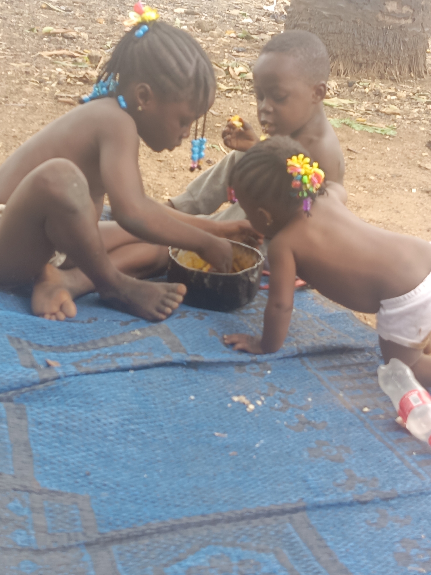 Three children are in a natural environment outdoors, apparently eating from a dark-colored bowl. One child is seated with their feet on the blue patterned mat, with hands in the bowl. A second child sits to the right of that child and appears to also be eating from the bowl. The third child is kneeling, reaching into the bowl. A water bottle lays on the ground near the kneeling child. The location is identified as Akwanga, Nigeria.