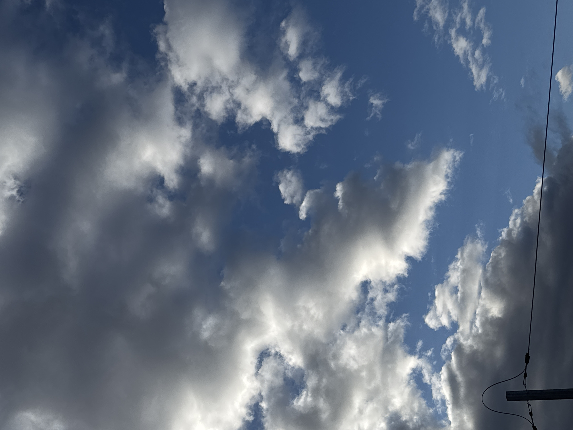 The image displays a dynamic sky dominated by clouds of varying densities and colors against a blue backdrop. The overall scene shows a mixture of bright white, fluffy cumulus clouds, and darker, denser grey clouds, indicating a partially cloudy condition with significant cloud cover.
On the left side of the frame, large, dark grey clouds are prominent, obscuring much of the blue sky. Moving towards the center and right, patches of deep blue sky become visible between scattered, brighter white clouds. Some of these white clouds appear more diffused and wispy, while others retain distinct, rounded shapes. The sunlight illuminates the edges and tops of the white clouds, creating a strong contrast with the darker, shadowed areas of the denser clouds.
On the far right edge of the image, a thin, dark wire extends diagonally from the top towards the bottom. Below this wire, a small portion of a metallic, horizontal structure, possibly a pole or support, is visible.
There are no people or discernible activities taking place. The time of day appears to be daytime, likely late morning or afternoon, given the clear blue sky patches and the evident sunlight on the clouds. The weather is dynamic, characterized by a mix of cloud types and densities. No specific location cues are visible within the frame beyond the generic utility wire and structure, typical of many developed areas, with the location context being Oktyabrsky, Russia. There is no visible text in the image.