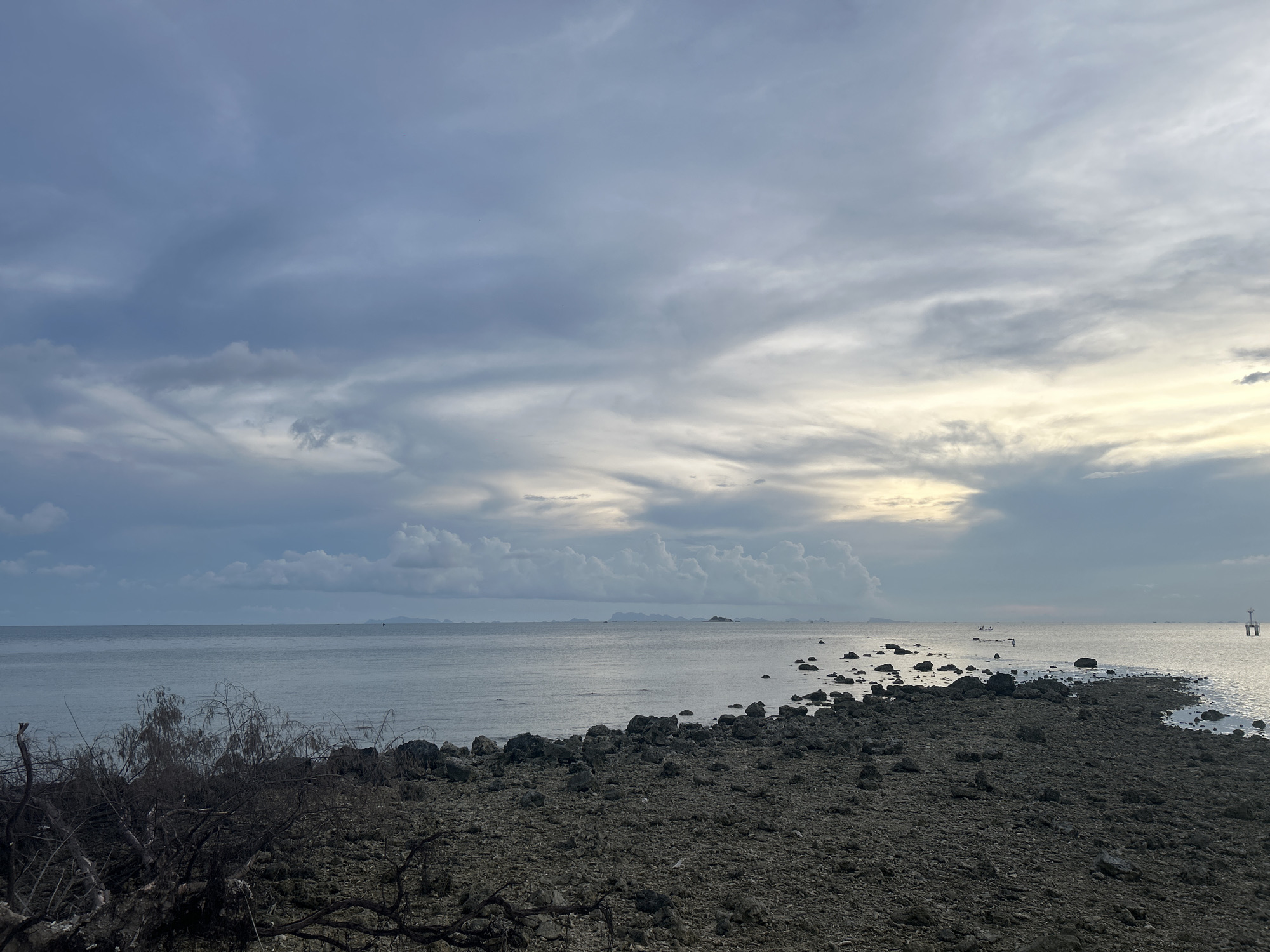 The image depicts a rocky shoreline at low tide under a cloudy sky in Baan Sri Thanu, Thailand. The sky is predominantly gray with patches of brighter, lighter clouds near the horizon. The ocean is calm and gray. The foreground shows a rocky beach with sparse vegetation, including some dark, dry branches and shrubs on the left. The shoreline extends into the sea, appearing as a pathway of rocks and boulders. In the distance, there are small islands and what appears to be a boat or small structure on the water. A navigational marker is on the right side of the photo. There are no people visible in the image.