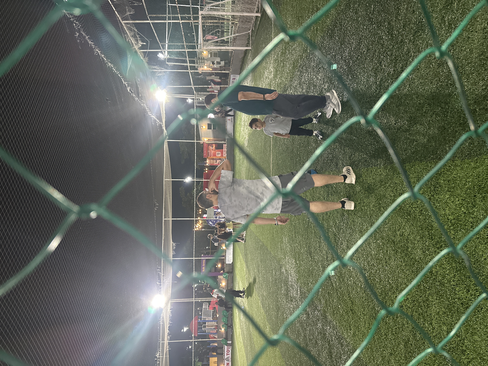 An exterior, medium shot captured at night reveals a green, artificial turf sports field with people present, viewed through a green chain-link fence. The field is illuminated by artificial lights, casting a glare on the wet-looking surface. In the foreground, a portion of the fence is visible, obscuring parts of the scene.
Several individuals are scattered across the field. On the right, a man wearing a dark t-shirt and dark pants stands near a young boy in a grey t-shirt and black pants. To the left of them, a man in a grey camouflage t-shirt and grey shorts faces away from the camera. In the middle ground, more people are seen, including a man in a white shirt and red collar shirt, and a group of individuals, some appearing to be women, further in the distance.
In the background, there are structures that suggest an entertainment or recreational area, with colorful playground equipment on the left. A sign that reads "Kids Arena" is visible above the playground. Soccer goals are present on the far right of the field. The overall impression is that of a public space, possibly a sports complex or recreational park, in Abuja, Nigeria, with activities taking place after dark.