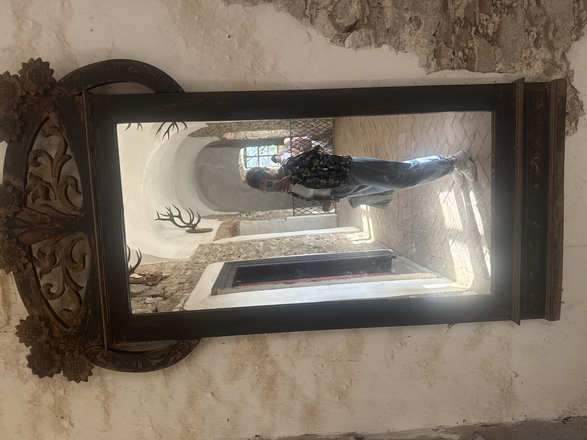 A person is reflected in an antique mirror in Chynadiiovo, Ukraine. The mirror has an ornate dark wood frame with sunflower carvings on the left side. The reflection shows a hallway with a vaulted ceiling, rough stone walls, and a window. Deer antlers are mounted on the wall. The person is holding a phone, presumably taking the photo. The walls around the mirror are aged and textured.