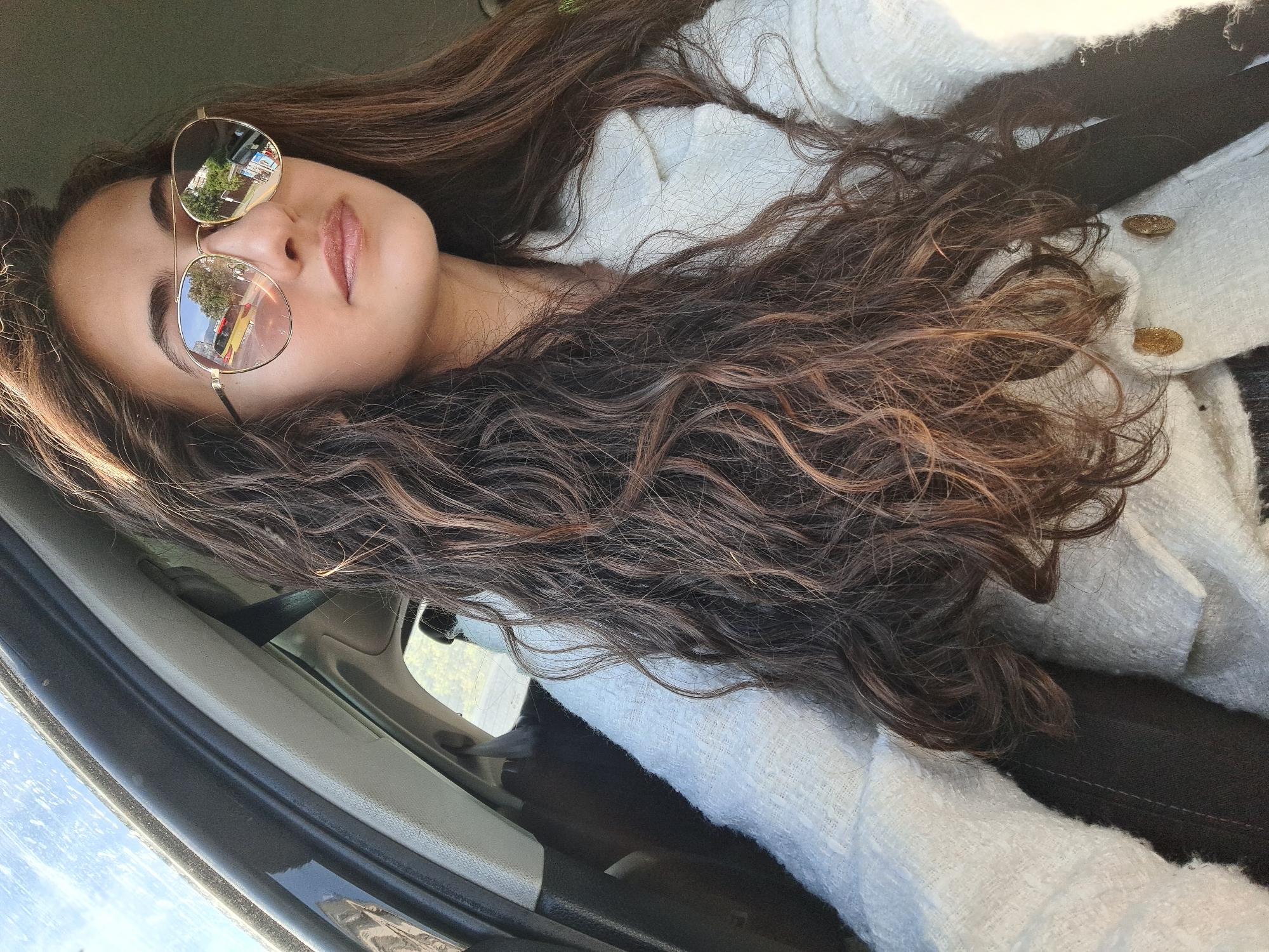 The image depicts a woman wearing sunglasses inside a car. The glasses reflect an outdoor scene with trees and buildings. The woman is wearing a white jacket with gold buttons and a black seatbelt. The car interior is visible, including the door frame and window. The city of Sofia, Bulgaria, is not discernible from the image.