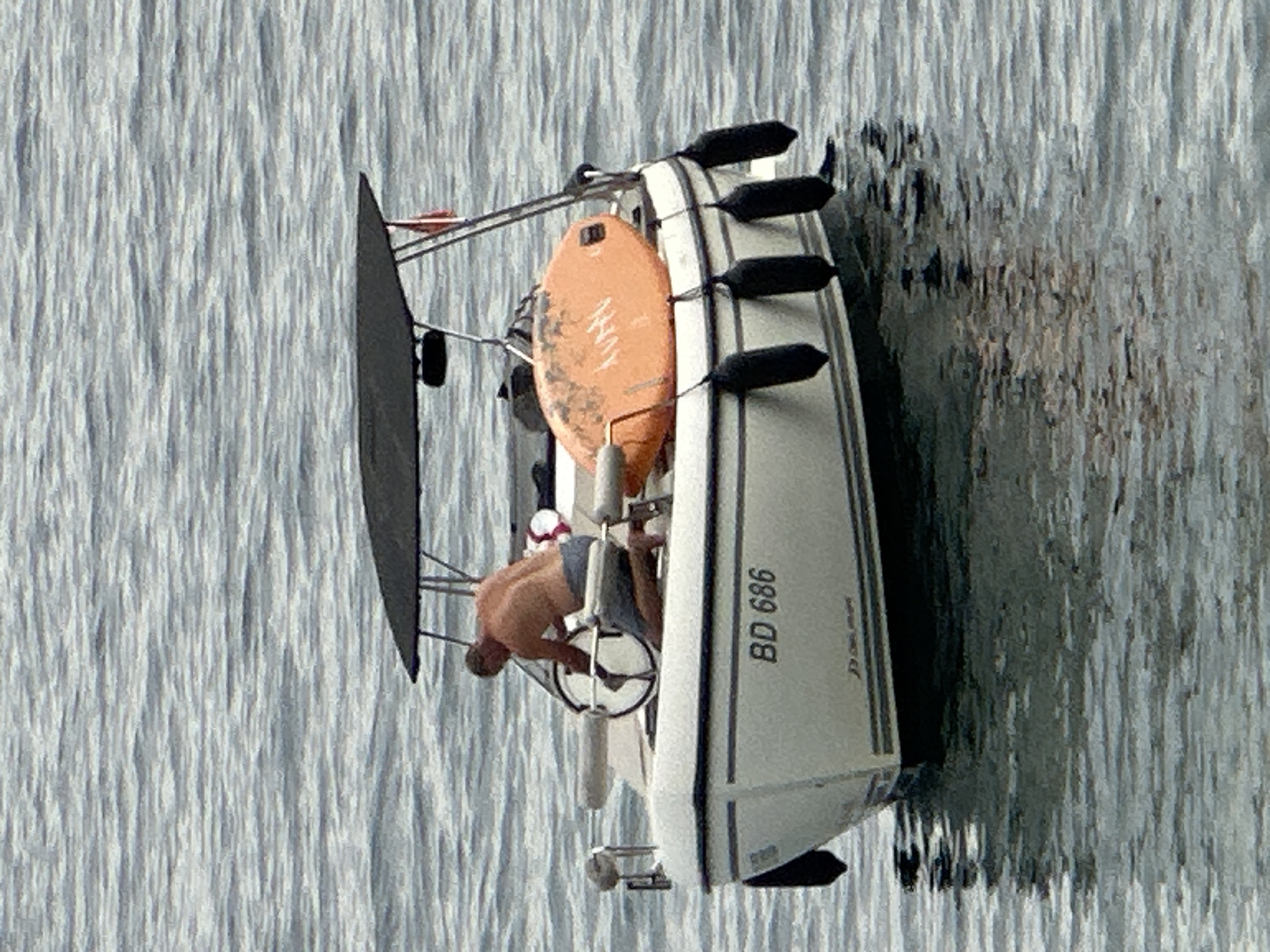 A man is kneeling on a white boat in calm, grey water. The man, who is shirtless and wearing grey shorts, is leaning forward and appears to be holding onto a steering wheel. The boat has a black canopy overhead supported by metal poles. An orange, oval-shaped object, possibly a life raft or surfboard, is secured to the boat. Several black fenders hang from the side of the boat. The boat has a registration number "BD 686" visible on its hull. The scene appears to be in the open water near Budva, Montenegro, judging by the boat's registration.