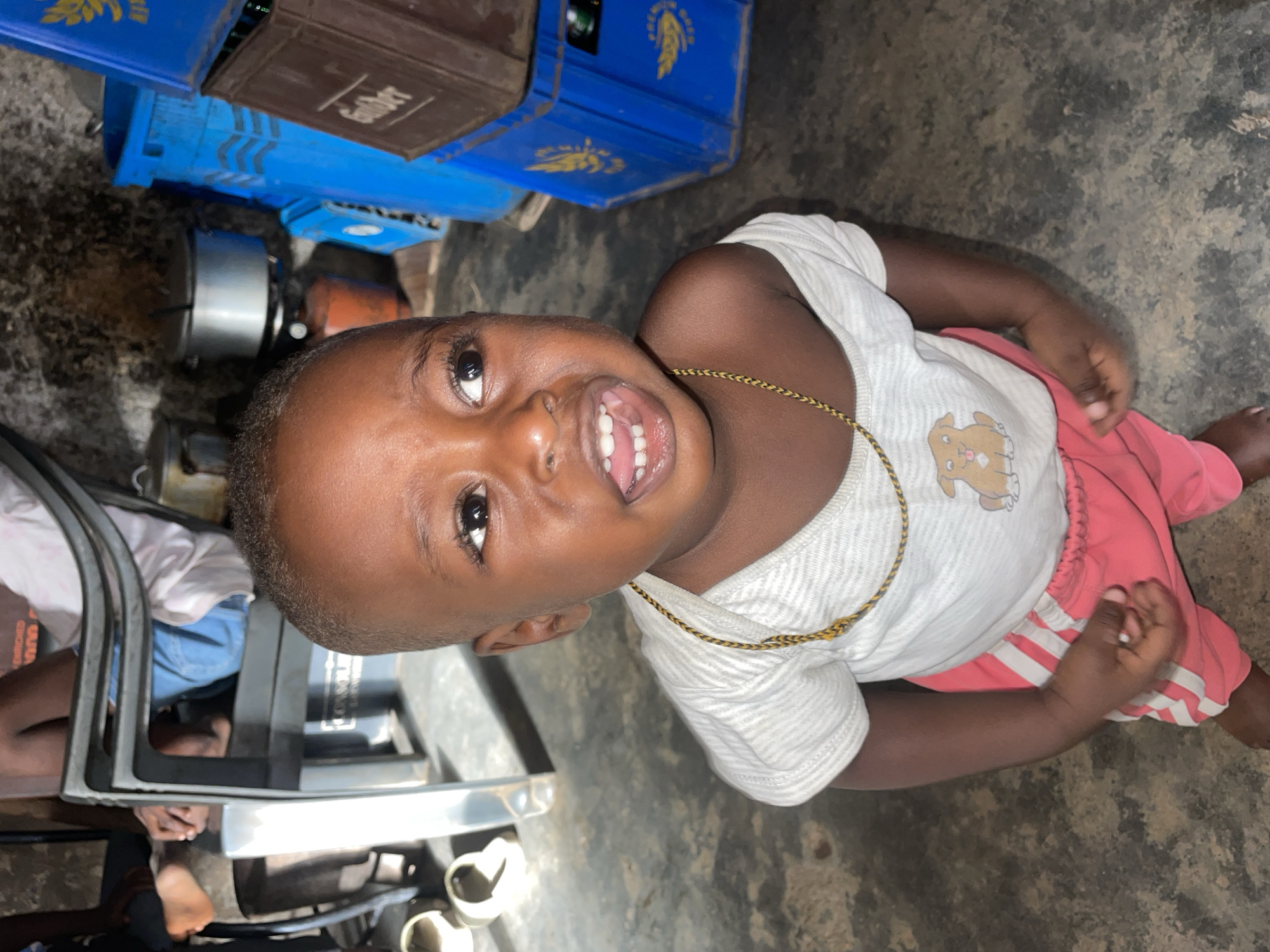 A toddler with dark skin, wearing a white T-shirt with a cartoon dog print and pink striped pants, looks up at the camera and smiles, revealing their teeth. The child wears a gold necklace. They appear to be sitting on the ground. In the background, there are blue and brown crates stacked on top of each other. A metallic folding chair is also visible. The setting seems to be an outdoor or semi-outdoor space, potentially in Jos, Nigeria.