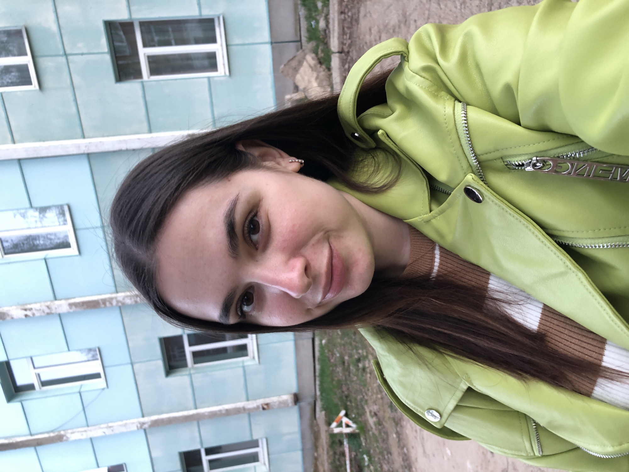 A woman in a light green jacket takes a selfie. Her long, dark hair is parted in the middle. She is wearing a brown and white striped sweater underneath the jacket. The background shows a multi-story building with multiple windows, suggestive of residential housing. The location is Noviy, Russia, as indicated by the prompt. The ground appears to be a combination of concrete and grass.