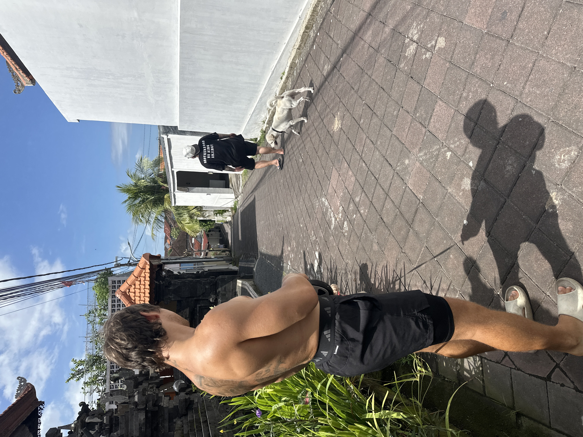 The image, shot in a residential area in North Kuta, Indonesia, shows a sunny outdoor scene. In the foreground, a shirtless person wearing black shorts and white sandals stands near green foliage, possibly a garden. In the middle ground, a person in a black t-shirt, shorts, and a white cap walks a white dog on a leash. A white wall and building are in the background. The pavement is brick. A shadow of the photographer is cast on the ground. Overhead, power lines are visible against a blue sky with scattered clouds. Balinese architecture with dark stone and brickwork elements is also present.