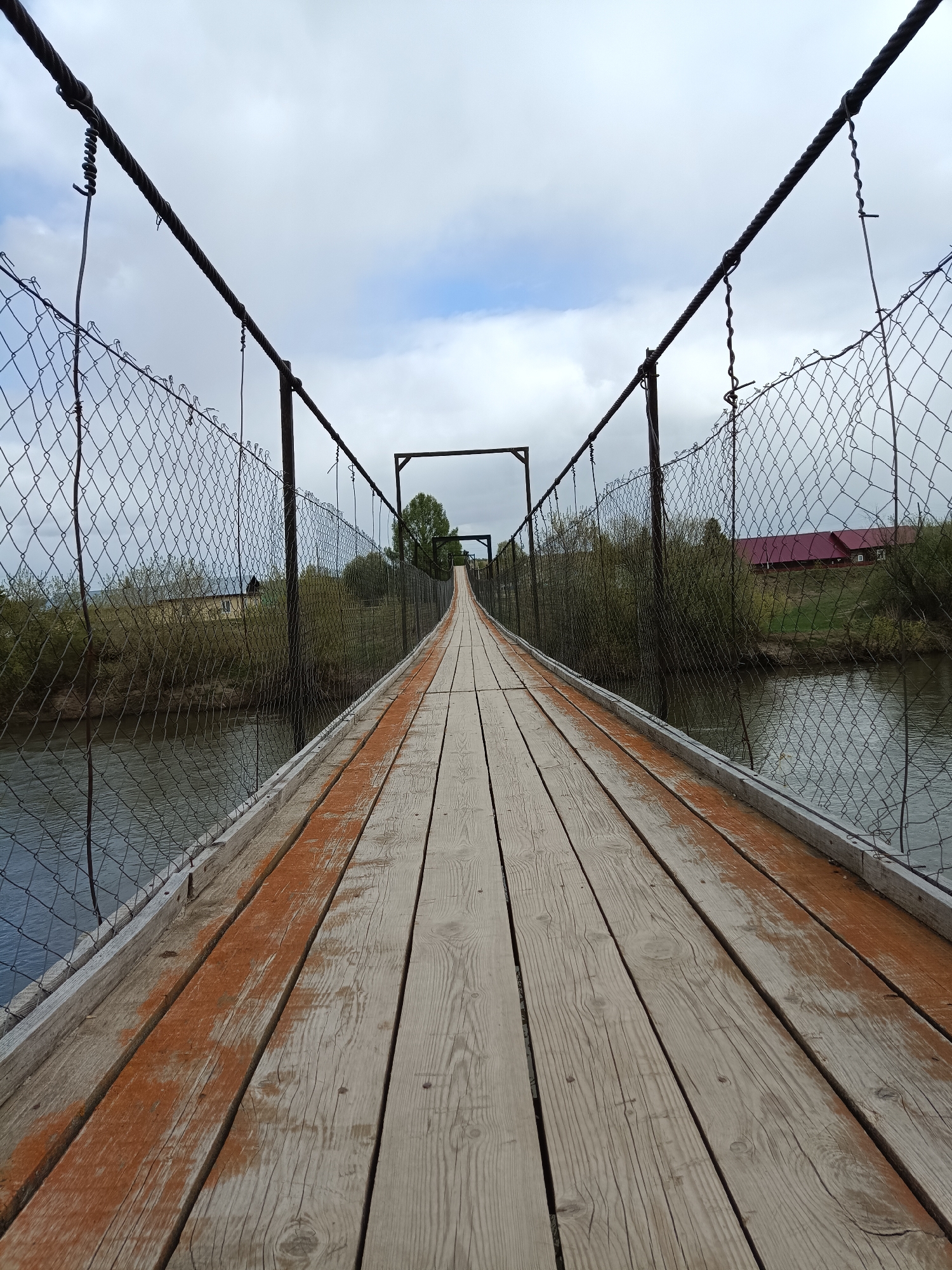 The image shows a long, narrow suspension bridge in Посад, Russia. The bridge has a wooden plank walkway with orange stains and is bordered by a wire fence on both sides. Heavy steel cables support the bridge's structure, running above the fencing and connecting to vertical metal posts. In the background, there is a body of water, likely a river or lake. On the far side of the water, there are trees, grass, and buildings including one with a red roof. The sky is overcast with grey and white clouds. There is no one visible on the bridge in the photograph.