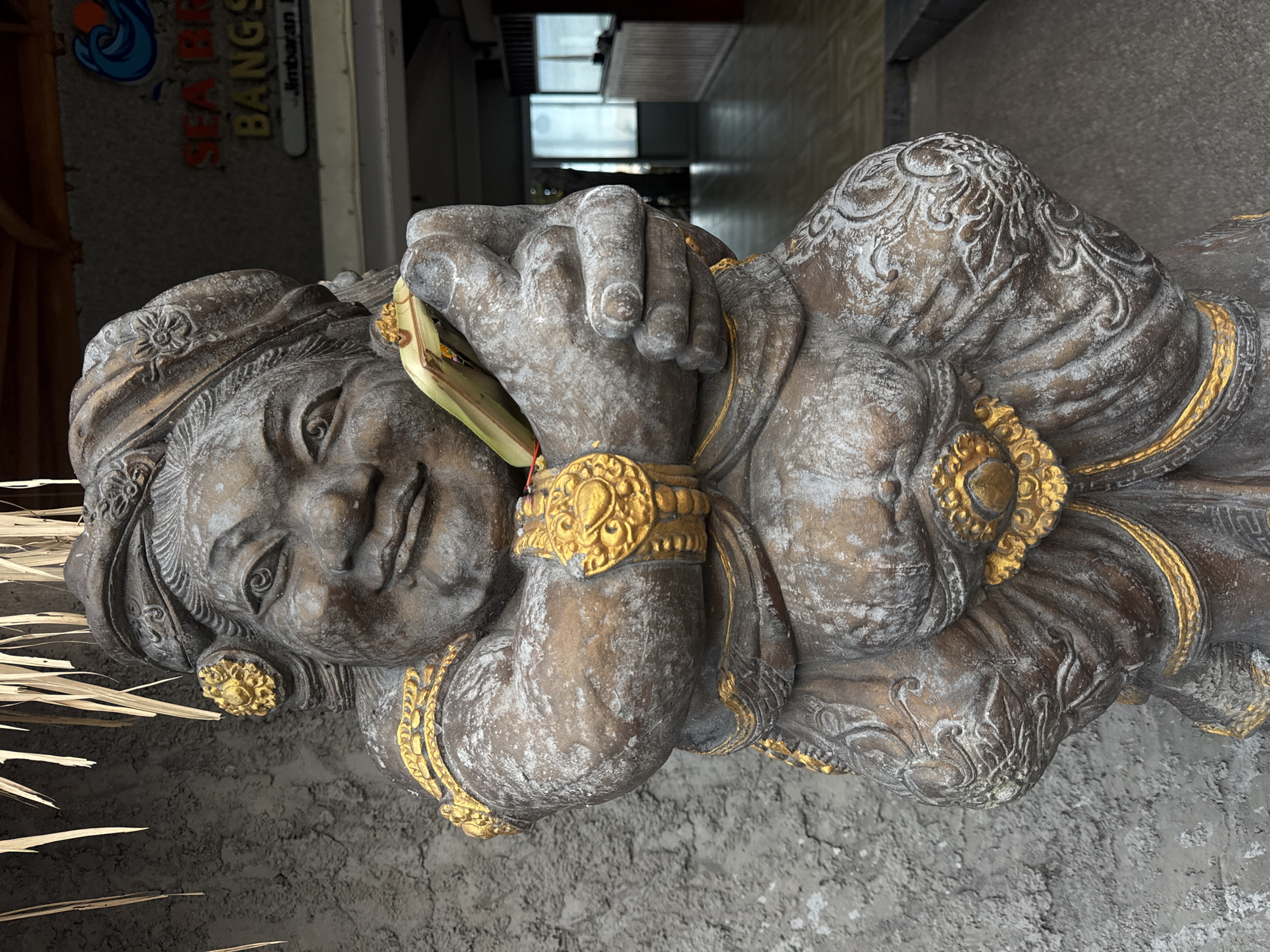 A weathered, ornate stone statue of a reclining figure, likely Balinese in style, dominates the foreground. The statue features gold-colored jewelry and embellishments. In the background, a sign reads "SEA BR BANGS" with "Jimbaran" printed below it, confirming the statue is in Jimbaran, a city near Kuta, Indonesia. The statue is positioned on a textured, concrete surface. A bundle of straw or dry plant material sits adjacent to the statue. The statue is photographed at a tilted angle. Beyond the statue, a glimpse of an interior area reveals a bright window.