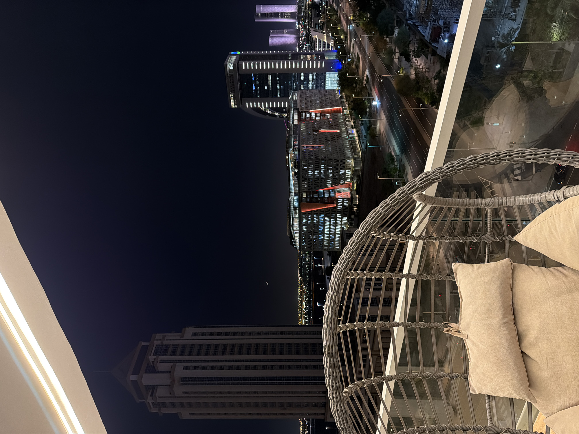 A balcony view at night showcases a cityscape with illuminated skyscrapers. In the foreground, a woven hanging chair with beige cushions faces the city. The balcony railing, made of glass and white metal, is visible. Beyond the railing, the city skyline features several modern buildings of varying heights, some with prominent lighting. A crescent moon is visible in the dark night sky. A street with cars and streetlights runs through the middle ground. Construction sites and some greenery are also present in the distance.