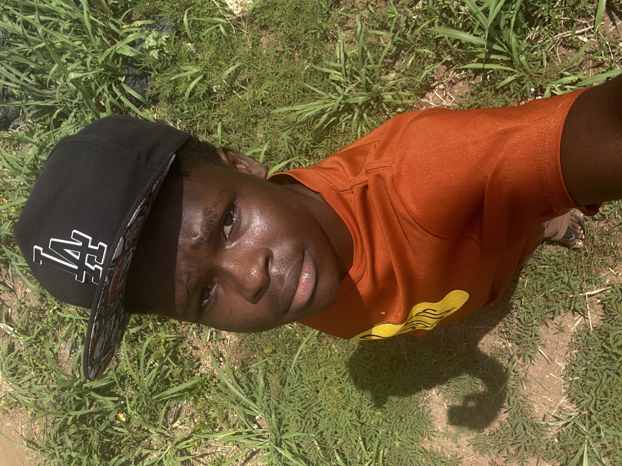 The image presents a head-on, high-angle portrait of a young person, rotated 90 degrees clockwise. The subject, who has dark skin, is looking directly at the camera with a neutral to slightly serene expression. Their skin glistens, suggesting exposure to sunlight.
They are wearing a black baseball cap adorned with a white "LA" logo on the front, and the cap's brim features a patterned underside. The person is also dressed in an orange t-shirt, which has a distinct yellow, wavy design located on its lower right side. A small portion of a sandal or flip-flop is visible near their right arm, indicating open footwear.
The setting is outdoors, with the ground predominantly covered in lush green grass, interspersed with areas of dry, light brown dirt. The strong shadows cast by the person on the ground clearly indicate bright, direct sunlight, suggesting the photograph was taken during the daytime under clear weather conditions. The overall environment appears natural and somewhat unkempt. The activity captured is a simple pose for the camera. Given the context, this scene is set in Ihiagwa, Nigeria.