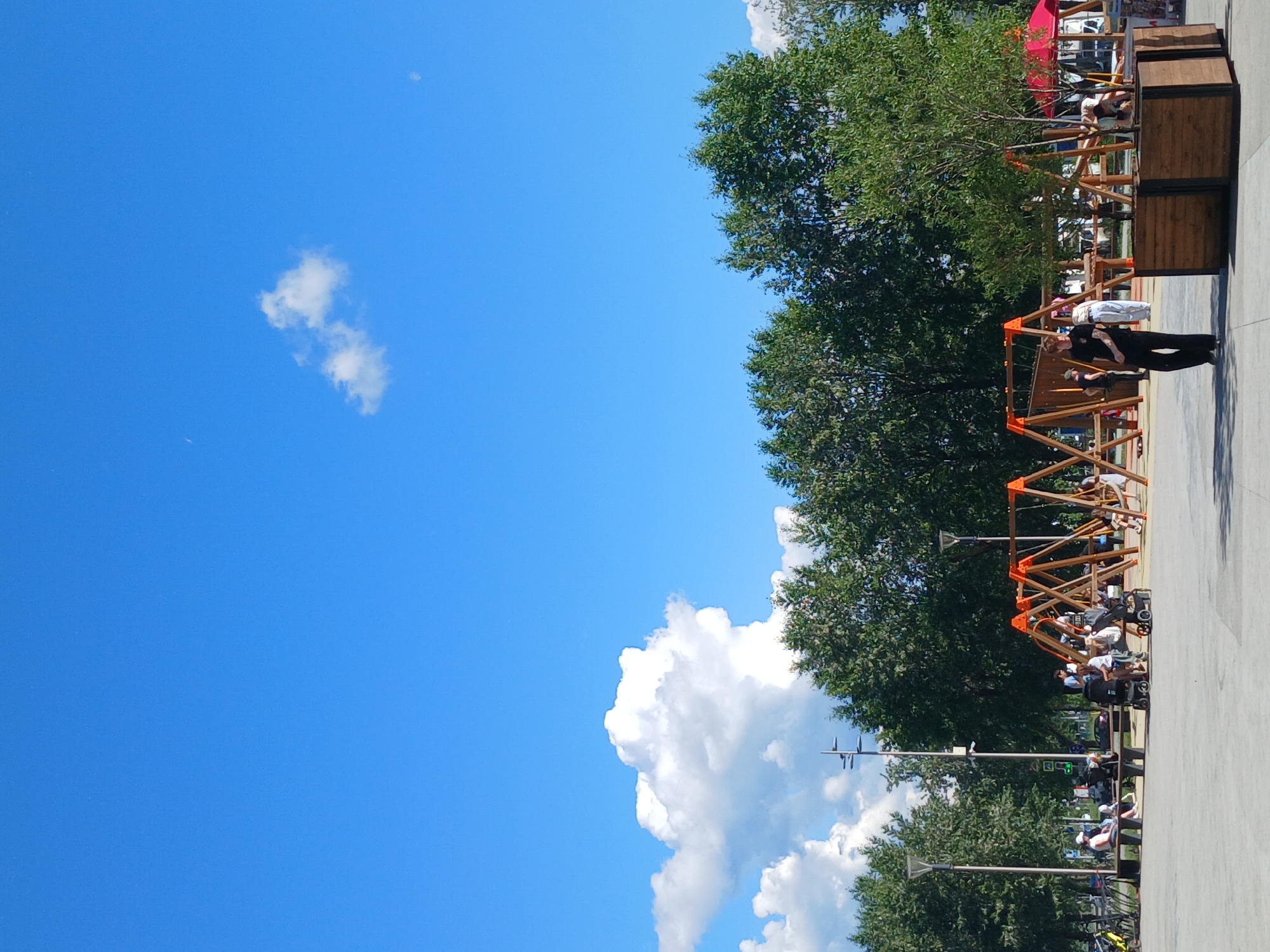 The image shows an outdoor public space in Yekaterinburg, Russia, on a sunny day with a bright blue sky and scattered white clouds. In the foreground, a paved area leads to a playground with swings. Several people are present, including adults sitting on benches and children playing on the swings. Some individuals are pushing strollers. In the background, there are mature trees, streetlights, and a red-roofed structure that may be part of the playground or a nearby facility. A person in a black shirt and pants is walking in the foreground. The scene appears to be a well-maintained park or recreational area.