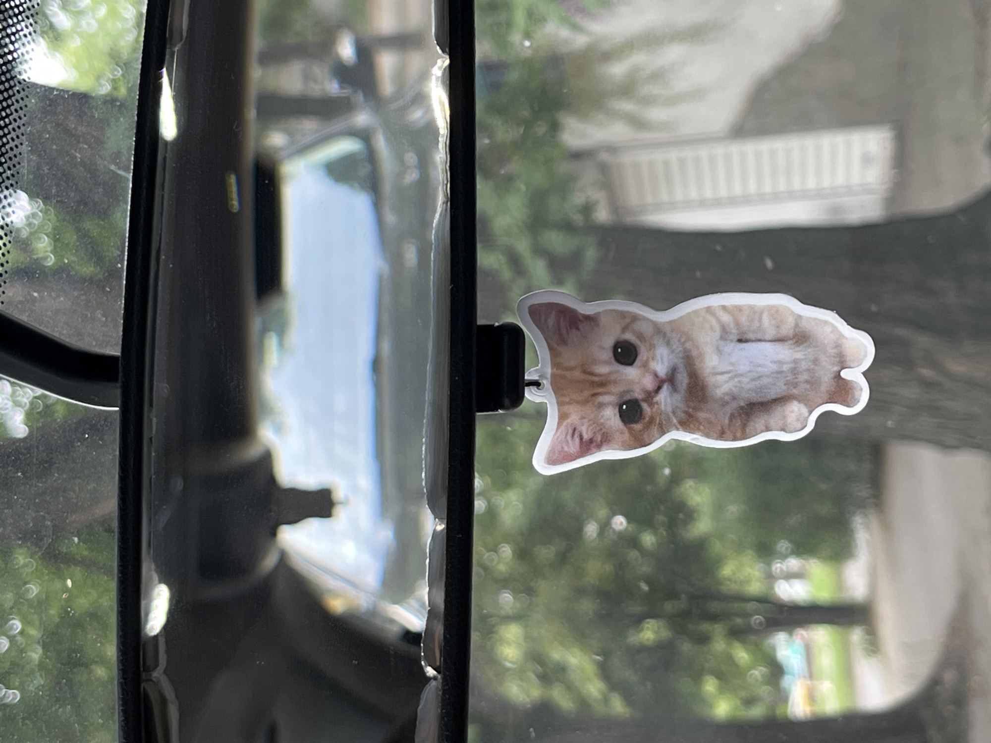An orange tabby kitten air freshener hangs from a car's rearview mirror. The kitten is depicted as a cartoonish sticker with large eyes and a pink nose. The background appears to be a residential street with trees and buildings, suggesting an outdoor setting in a city or town. The interior of the car is visible in the reflection of the mirror, showing a glimpse of the dashboard and windshield. The image was captured in Orizari, Bulgaria.