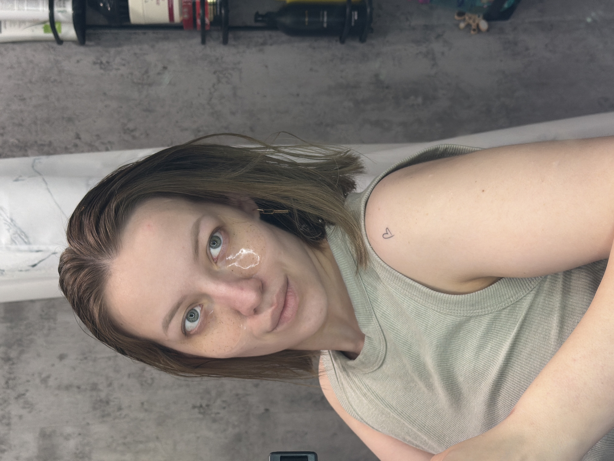 A woman with light brown hair, fair skin, and green eyes is pictured in a close-up shot. She has a translucent hydrogel patch under one eye and freckles on her face. She is wearing a light-colored ribbed tank top. A small heart tattoo is visible on her shoulder. The background includes a gray textured surface, a white patterned surface, and several bottles on a shelf. The location in the city of Reshetnikovo, Russia, cannot be determined from the image.