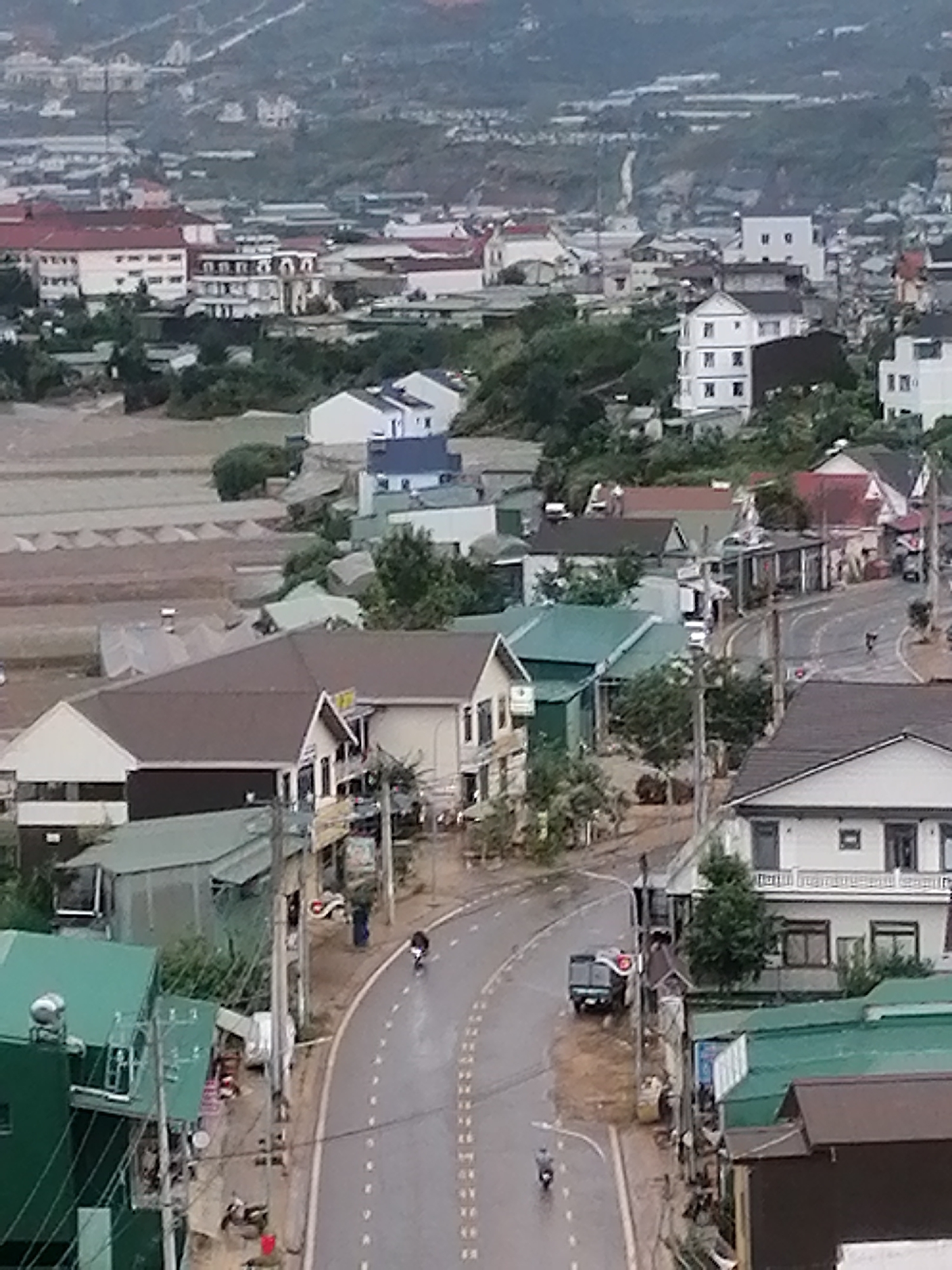This image depicts an elevated, high-angle view of a curving, wet asphalt road in Lạc Dương, Vietnam. The road is bordered by buildings and lush green foliage. Several vehicles are present, including a white van with a red light on top parked on the right side of the road, and a scooter with a rider heading away from the camera on the left side of the road. Another scooter is visible further up the road on the right. The buildings flanking the road are a mix of architectural styles, with some featuring brown, green, and white roofs. One building on the left has a prominent green roof and appears to be a commercial establishment. On the right side of the road, a white multi-story building with balconies is visible. In the background, a hillside covered with buildings and vegetation stretches into the distance, with some structures appearing to be greenhouses or agricultural buildings. The overall atmosphere suggests a moderately overcast or rainy day, given the wet road surface and muted lighting.