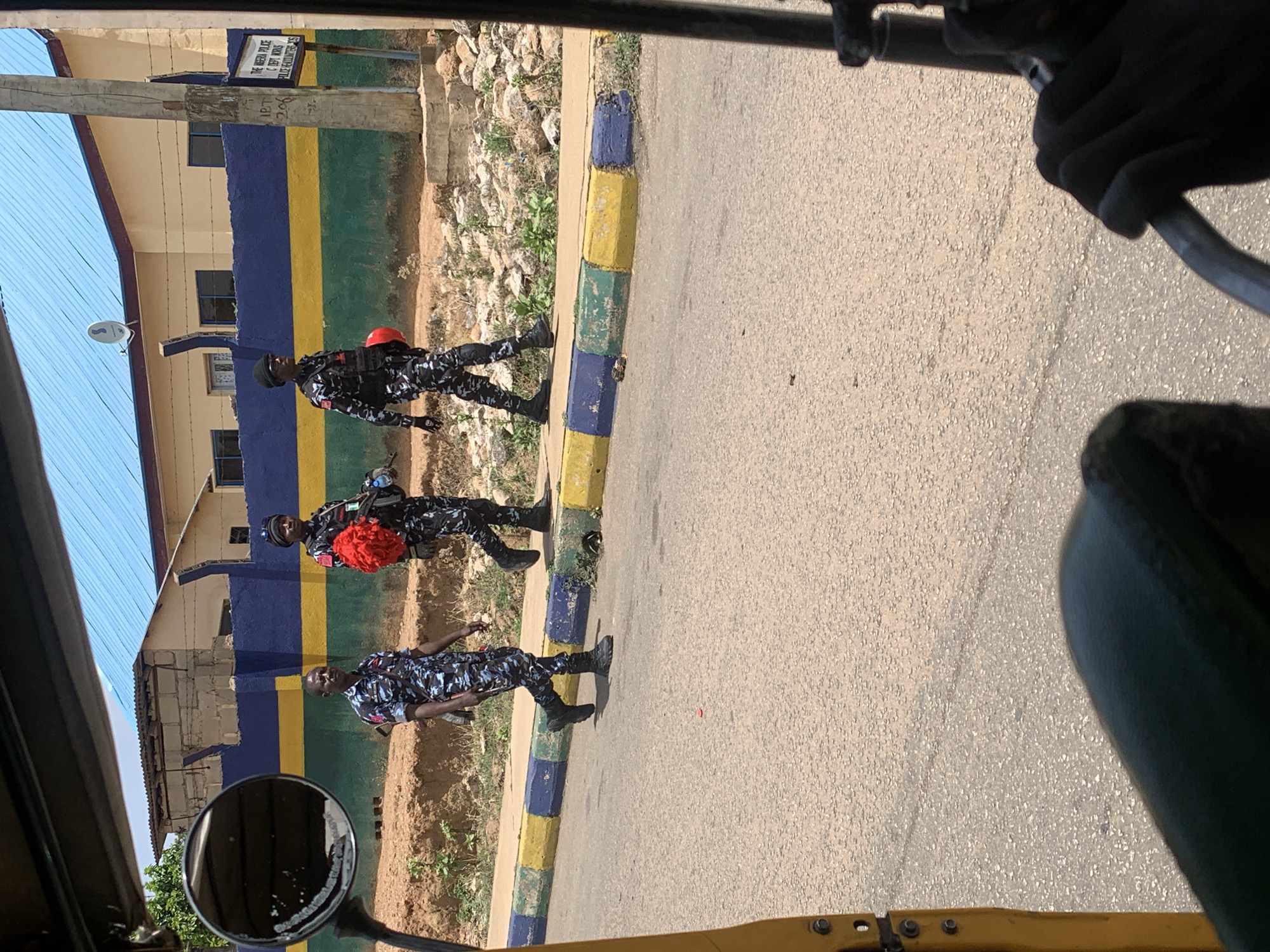 The image shows three armed policemen walking along a road in Jos, Nigeria. The policemen are wearing camouflage uniforms and black boots. Two of the policemen are carrying red equipment on their backs. The road has a curb painted with blue, yellow, and green stripes. A building with a blue, yellow, and green painted wall is visible in the background. The image is taken from inside a vehicle, possibly a motorcycle rickshaw (keke), showing parts of the vehicle's interior like the side mirror and handlebars. The setting appears to be a sunny day. The sign on the building indicates it is the Nigeria Police C Dept Works.